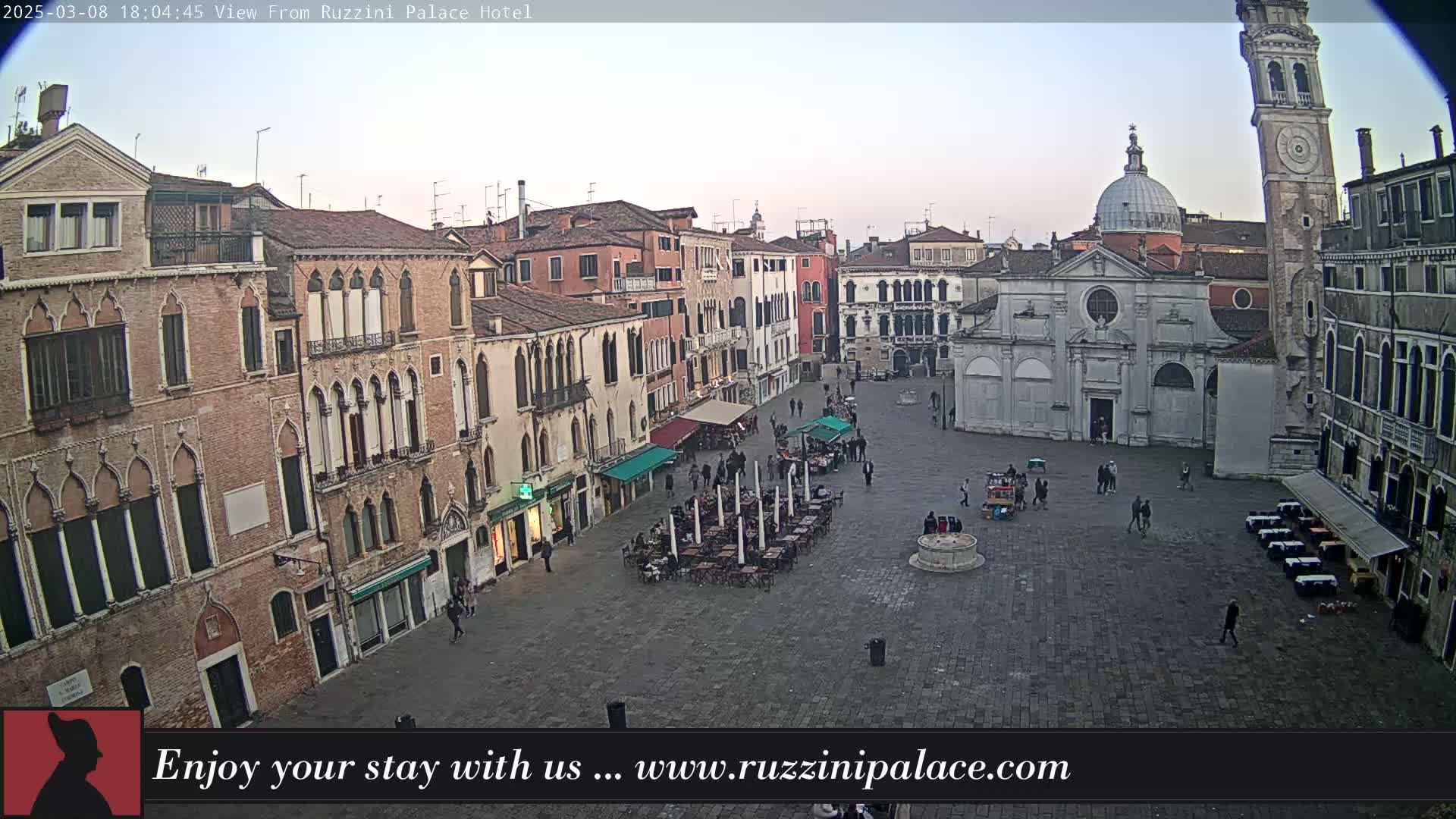 A twilight view of a large, mostly empty town square in Venice, surrounded by historic buildings, with a few people and outdoor seating areas visible.