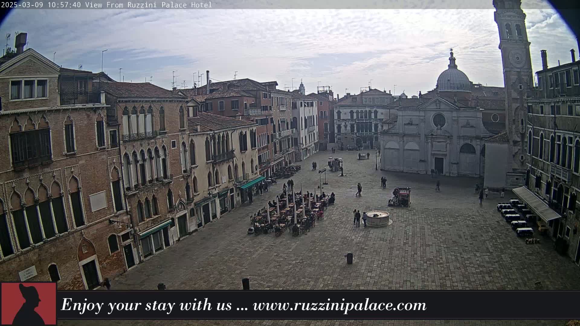 A mostly cloudy day reveals an expansive cobblestone square in Venice, Italy, surrounded by historic buildings, with several people and outdoor seating visible.
