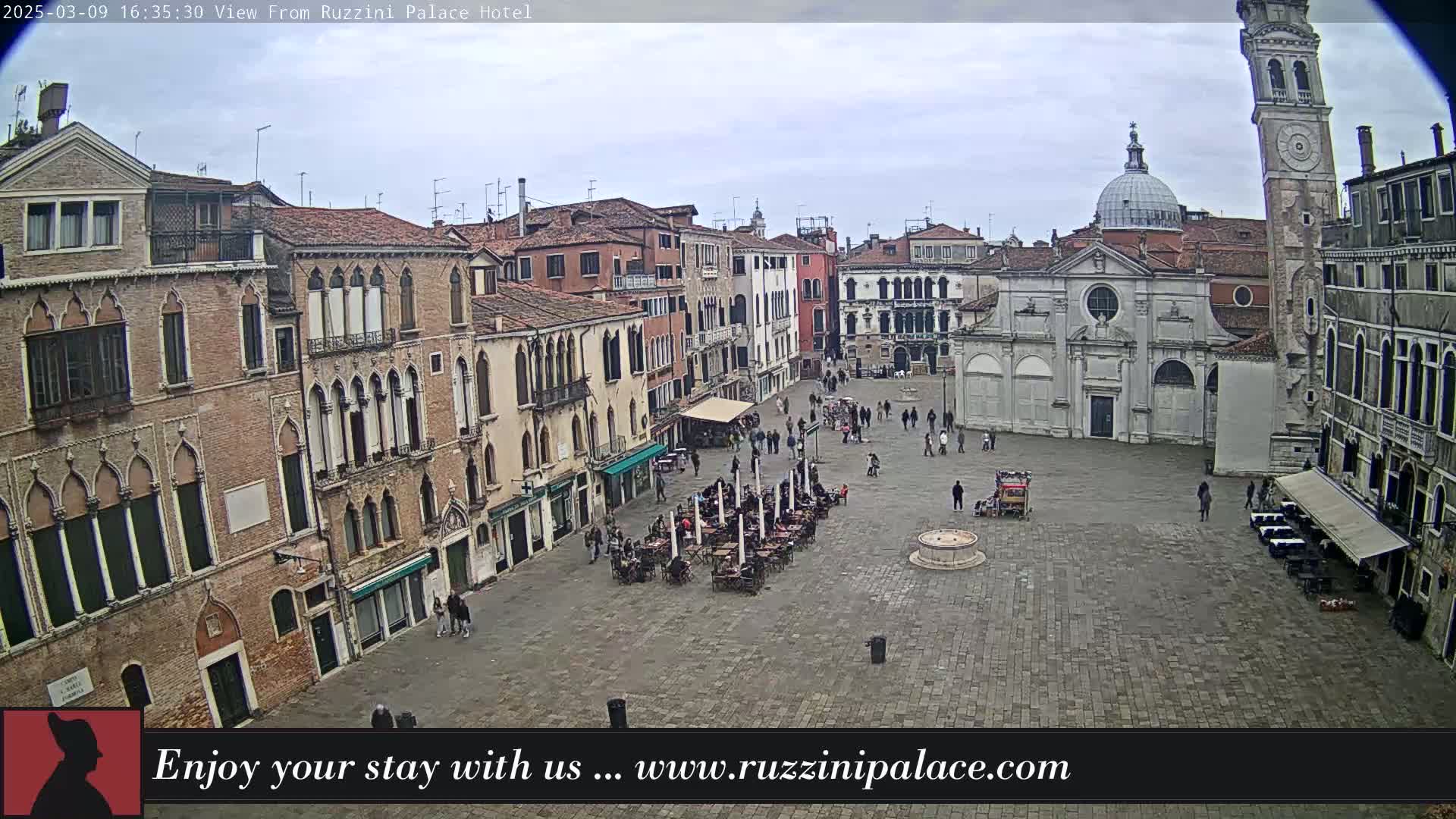 A large, paved square in Venice, Italy, is sparsely populated with people and outdoor seating, under an overcast sky.