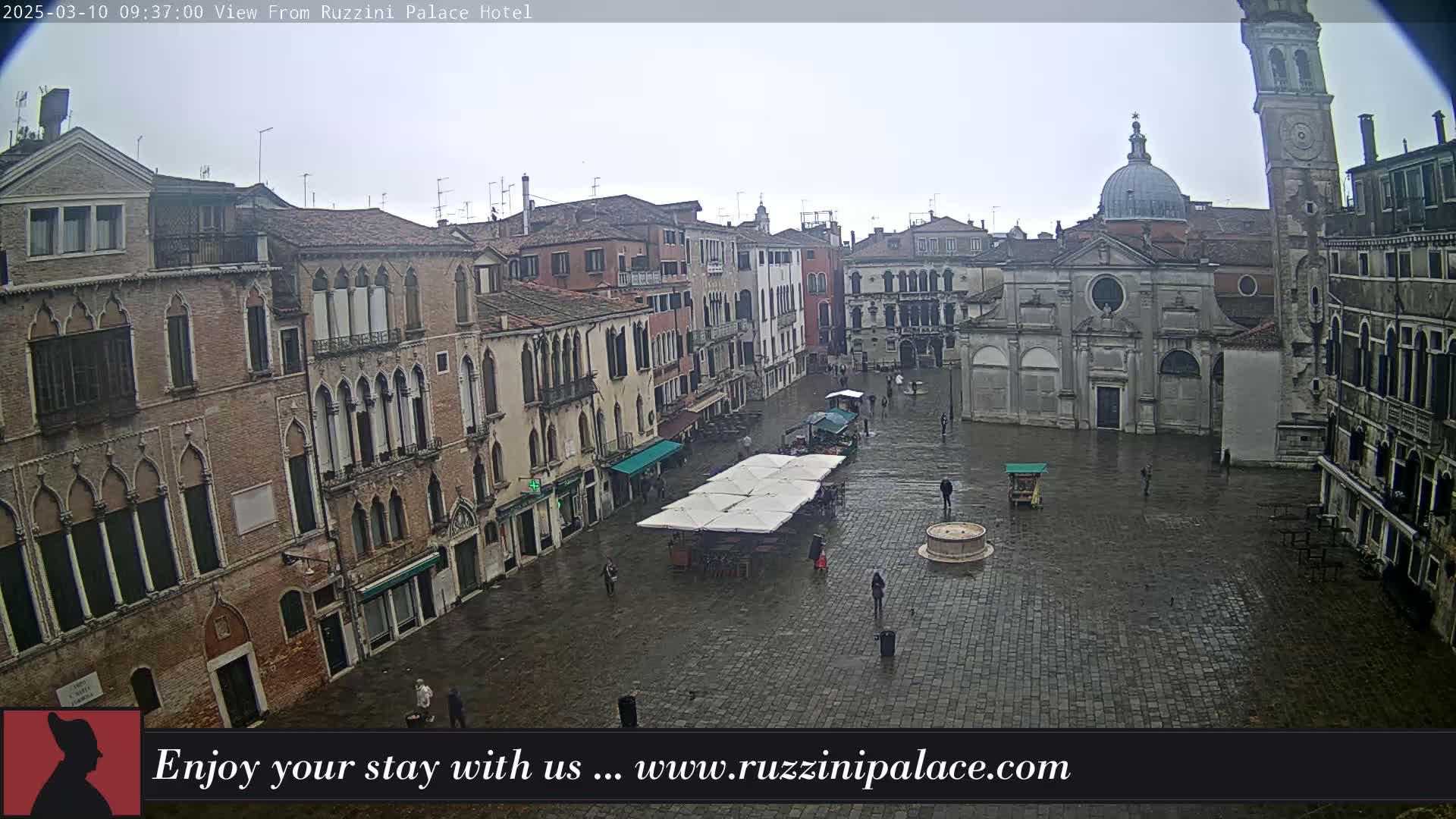 A wet, cobblestone square in Venice, Italy, is surrounded by old brick buildings, including a church and a tall tower, with a few people sparsely scattered throughout.