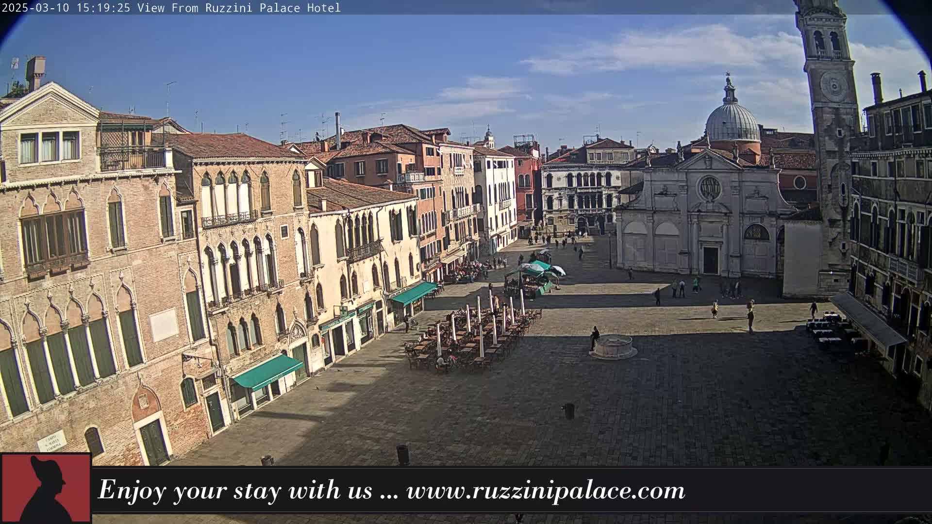 A sunny view of a large plaza in Venice, Italy, surrounded by old buildings, with a few people and outdoor seating visible.