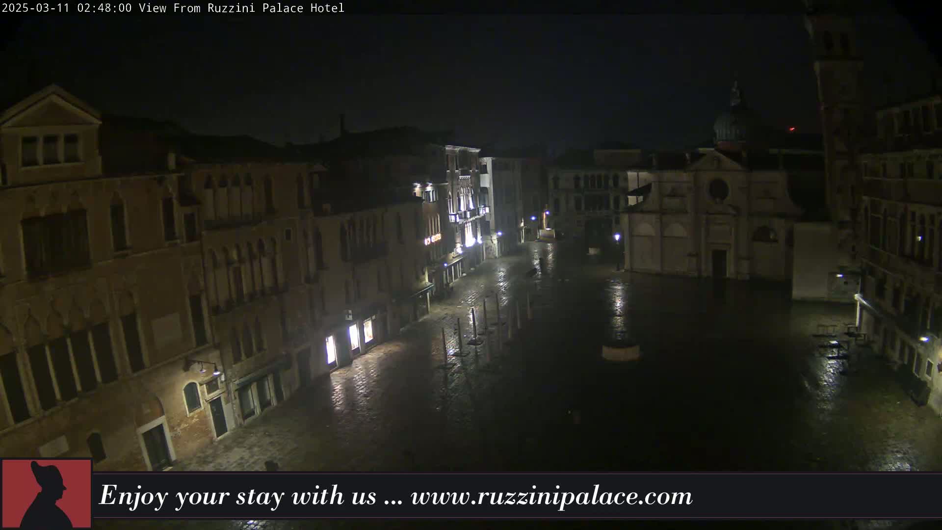 A rain-slicked, dimly lit European plaza at night is surrounded by old buildings.