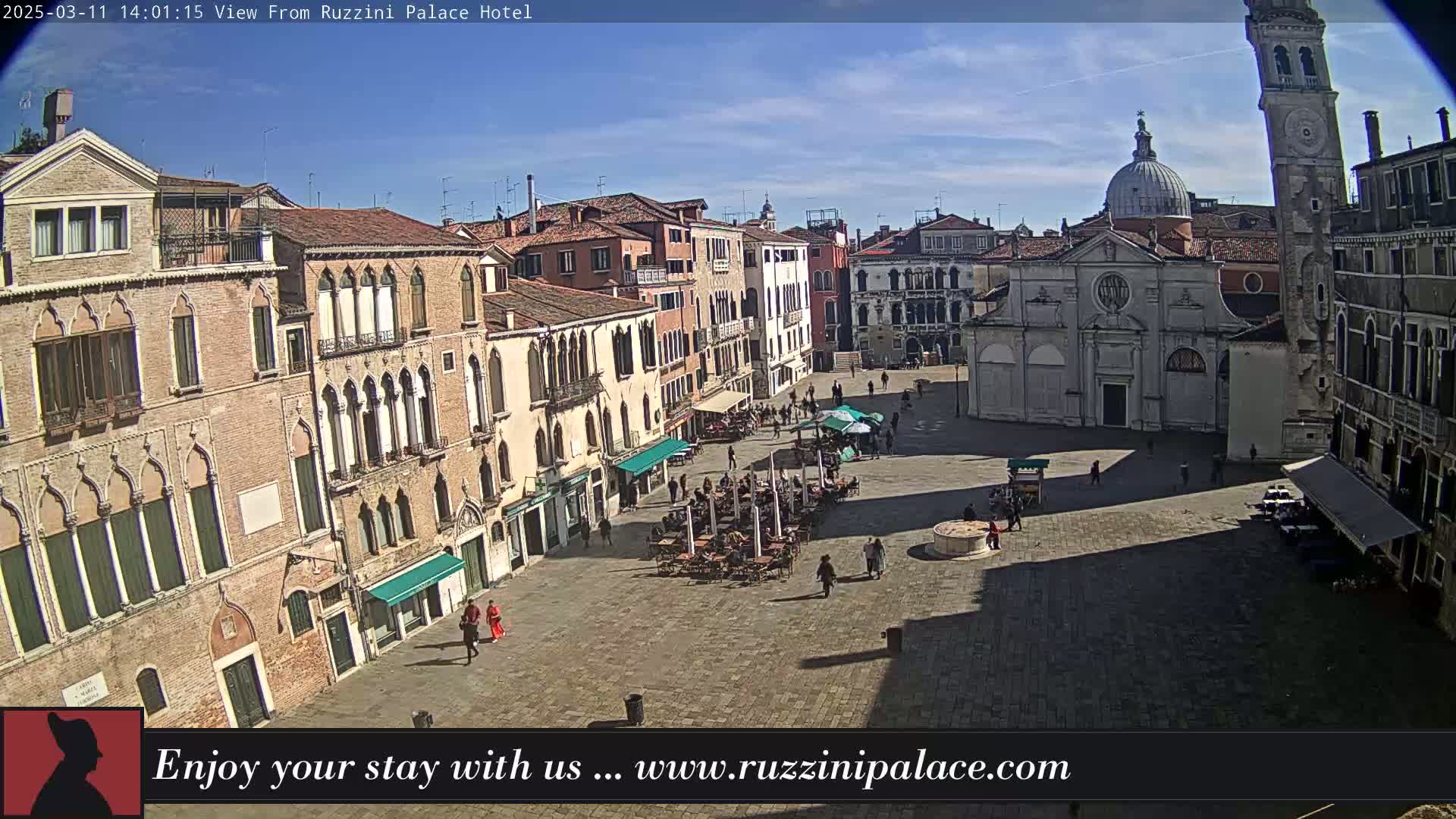 A sunny day reveals a large, paved square in Venice, Italy, surrounded by historic buildings, with people strolling and seated at outdoor cafes.