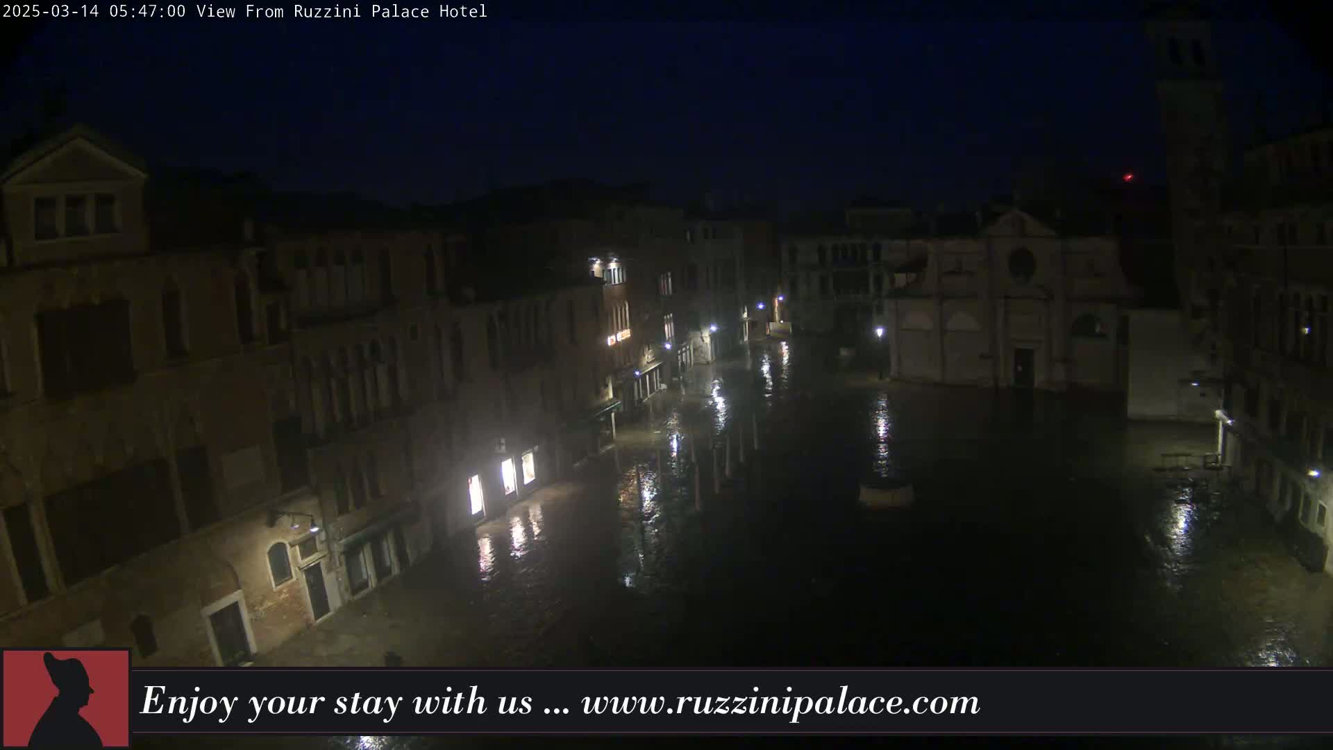 A nighttime, flooded plaza in Venice is surrounded by old buildings with illuminated windows under a dark sky.