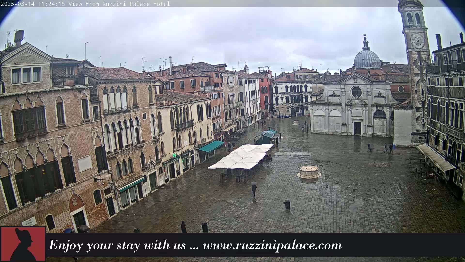 A rain-soaked cobblestone square in Venice, Italy, is surrounded by historic buildings, including a church with a dome and a tall clock tower.