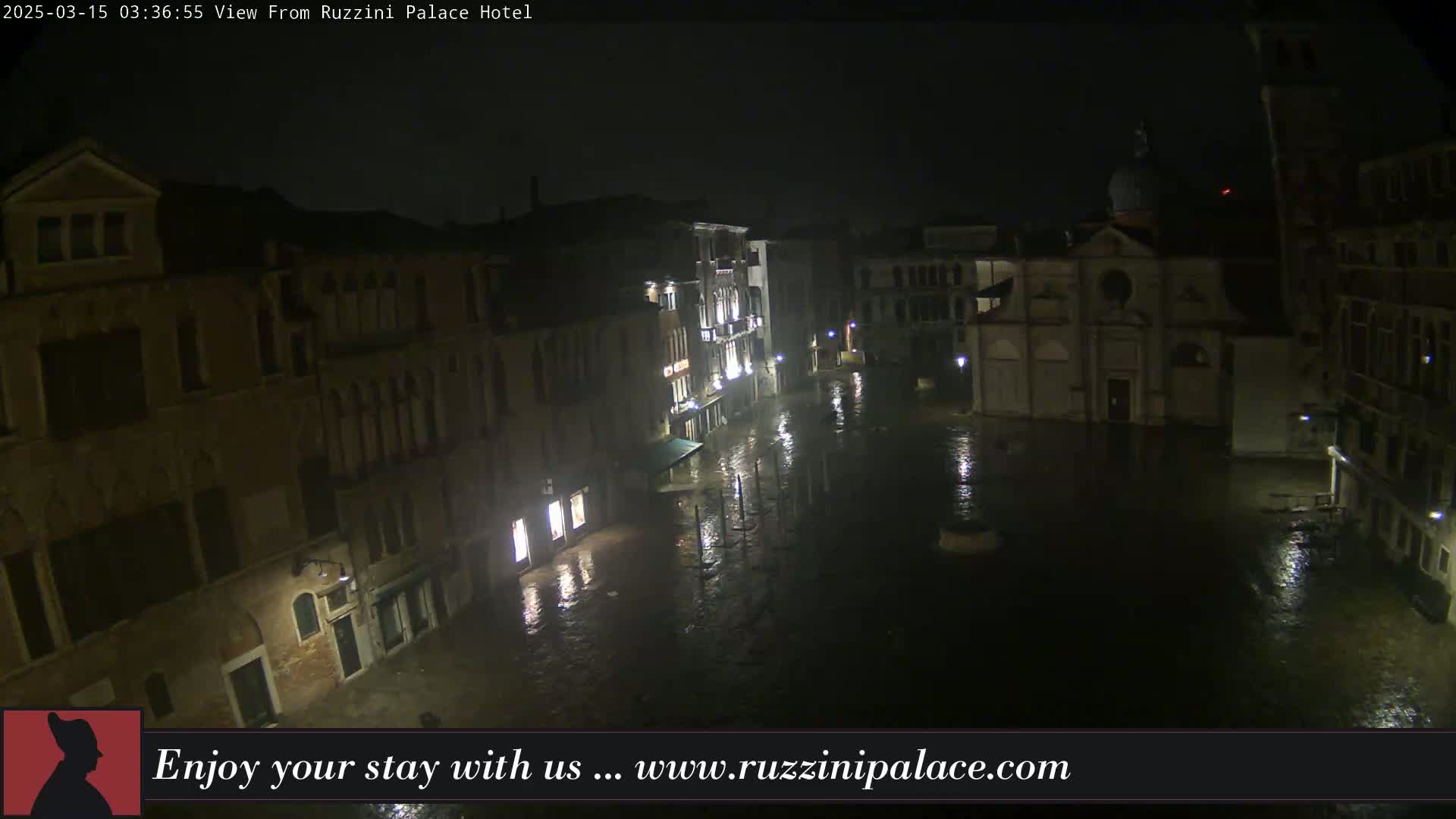 A nighttime view of a flooded plaza in Venice, surrounded by buildings, under dark and rainy conditions.