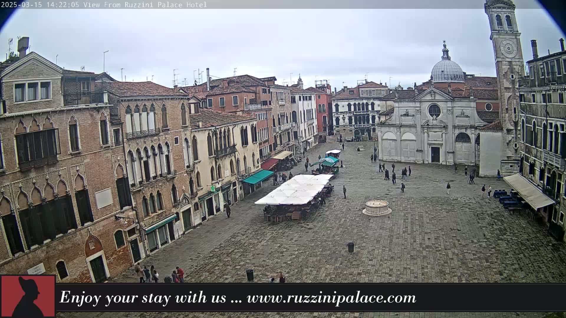 A large, cobblestone square in a European city is viewed from above on an overcast day, showing several buildings with arched windows, a church with a dome, a bell tower, and a few people walking around.