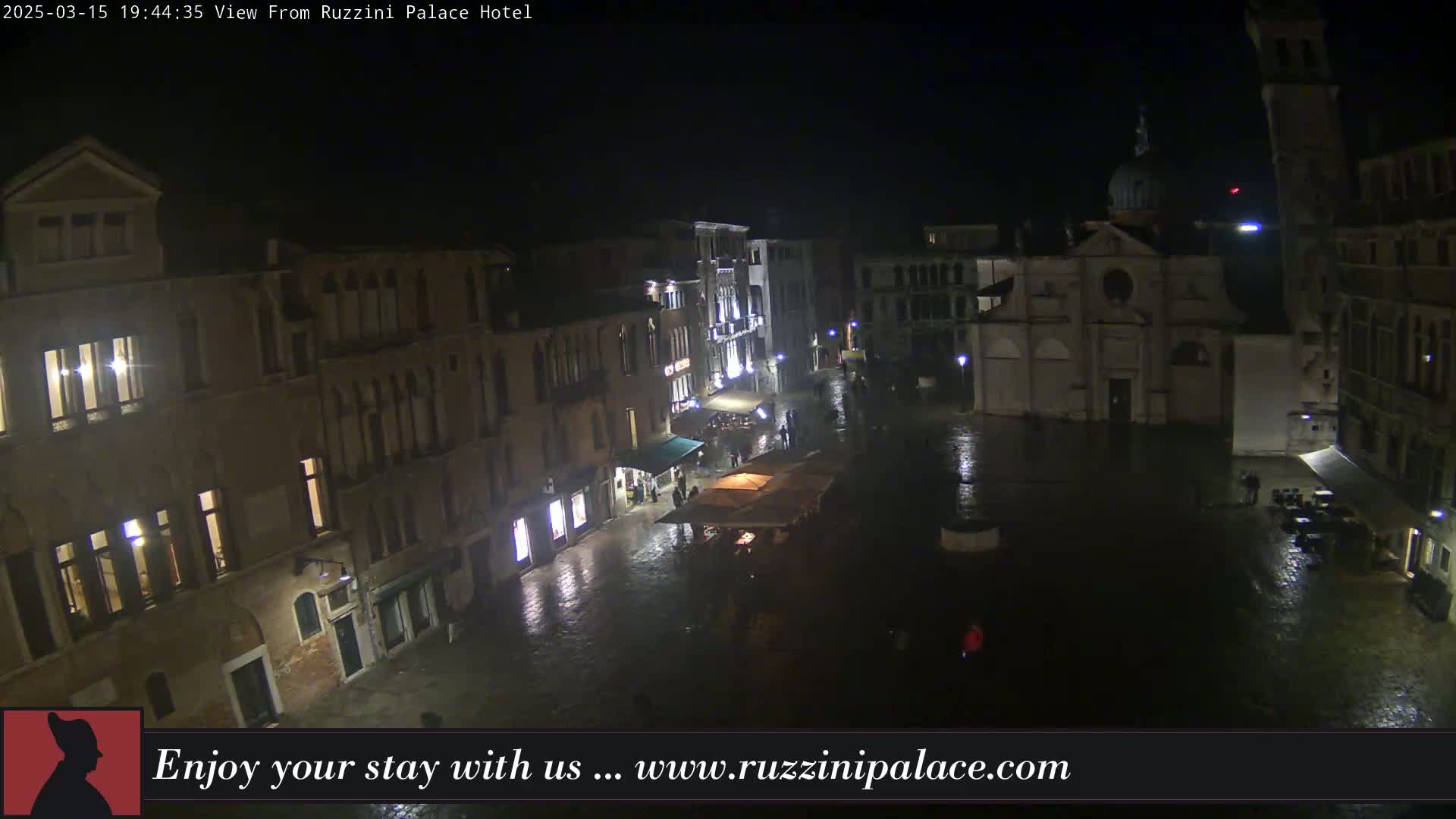 A nighttime, rain-slicked Venetian square is viewed from above, showing several buildings with illuminated windows and a few people sparsely scattered about.