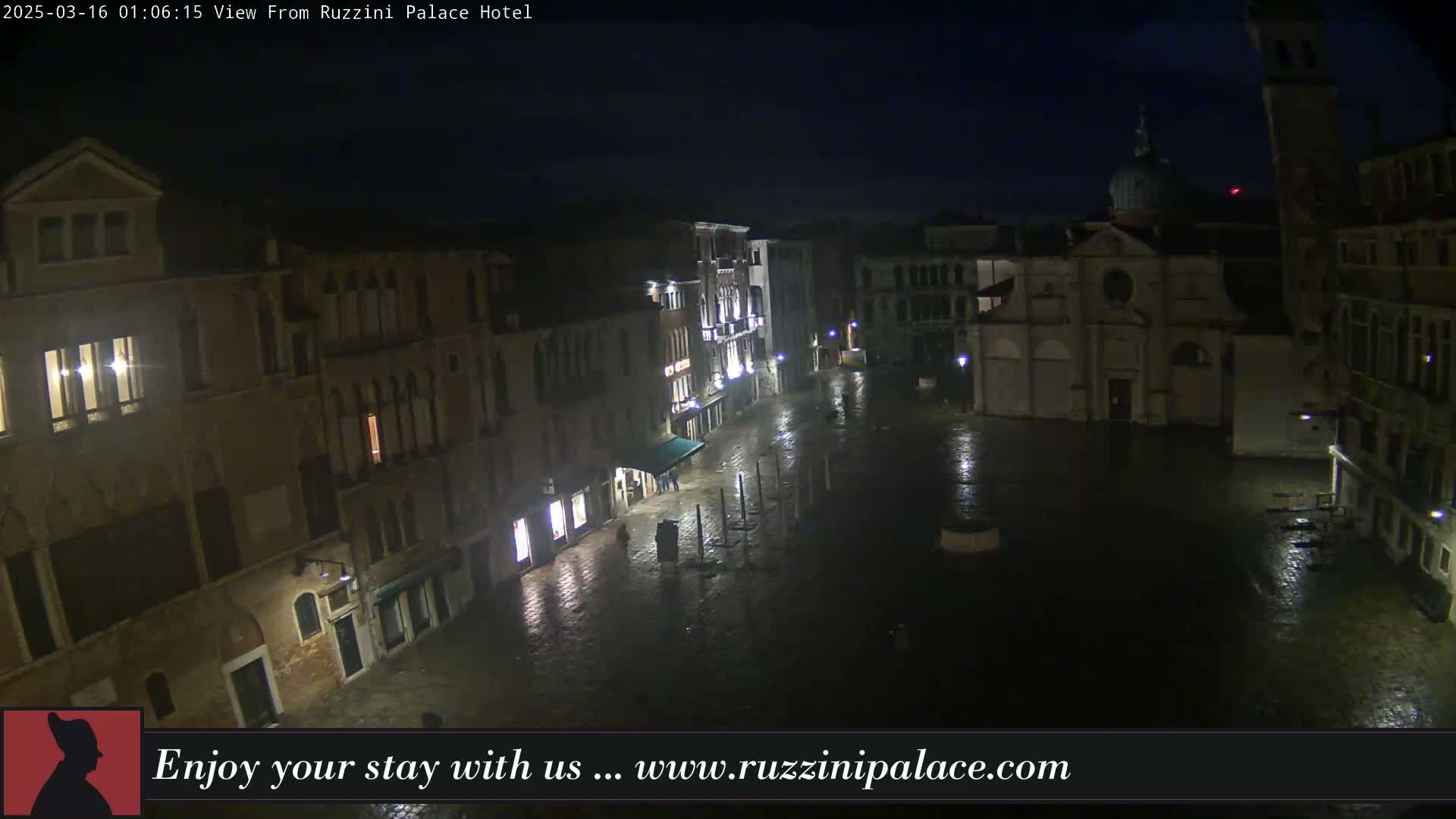 A nighttime, wet view of a large, mostly empty square in Venice, Italy, surrounded by old buildings and a church.