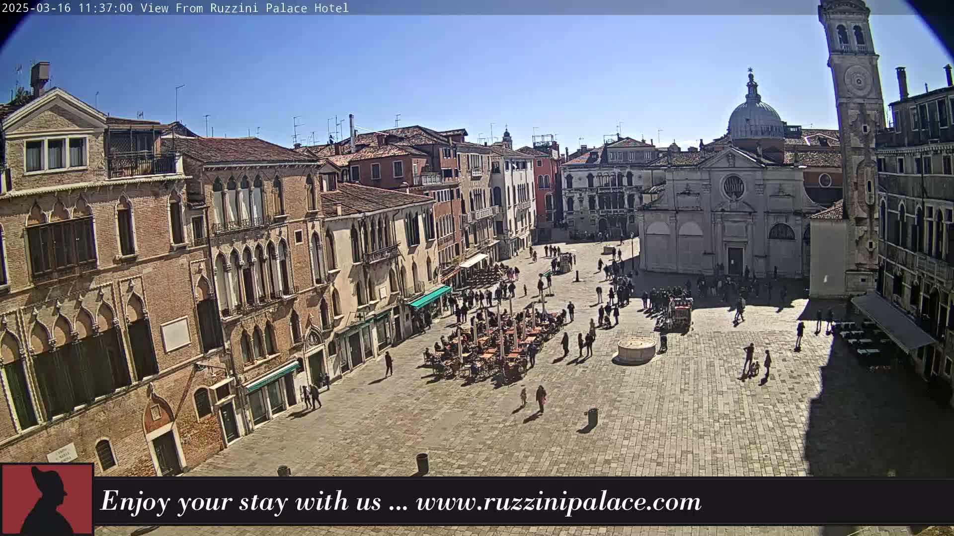 A sunny day reveals a large, paved square in Venice, Italy, surrounded by historic buildings, with people walking and sitting at outdoor cafes.