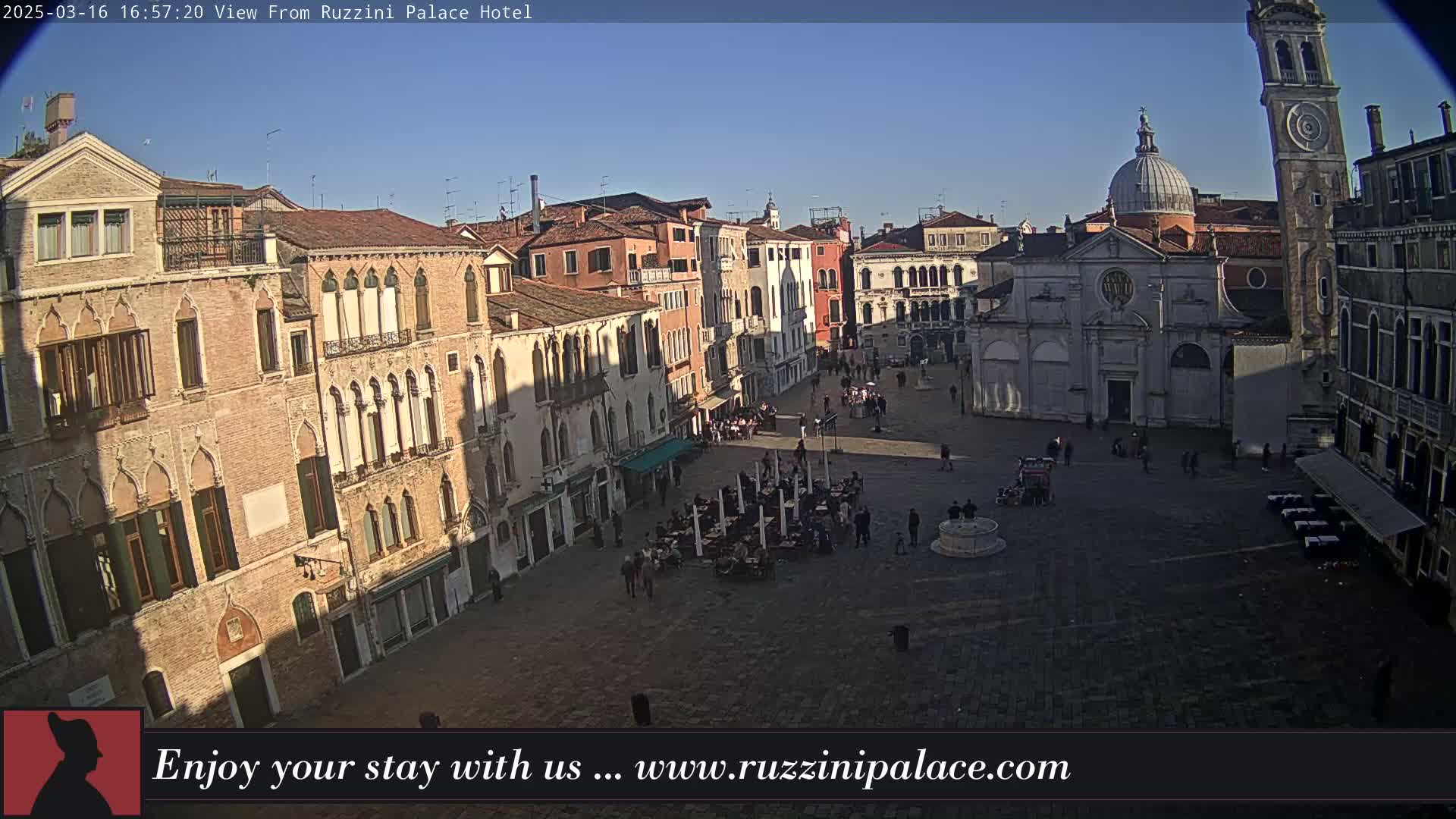 A sunny, high-angle view of a large, paved town square in Venice, Italy, surrounded by historic buildings, with a few people scattered throughout.