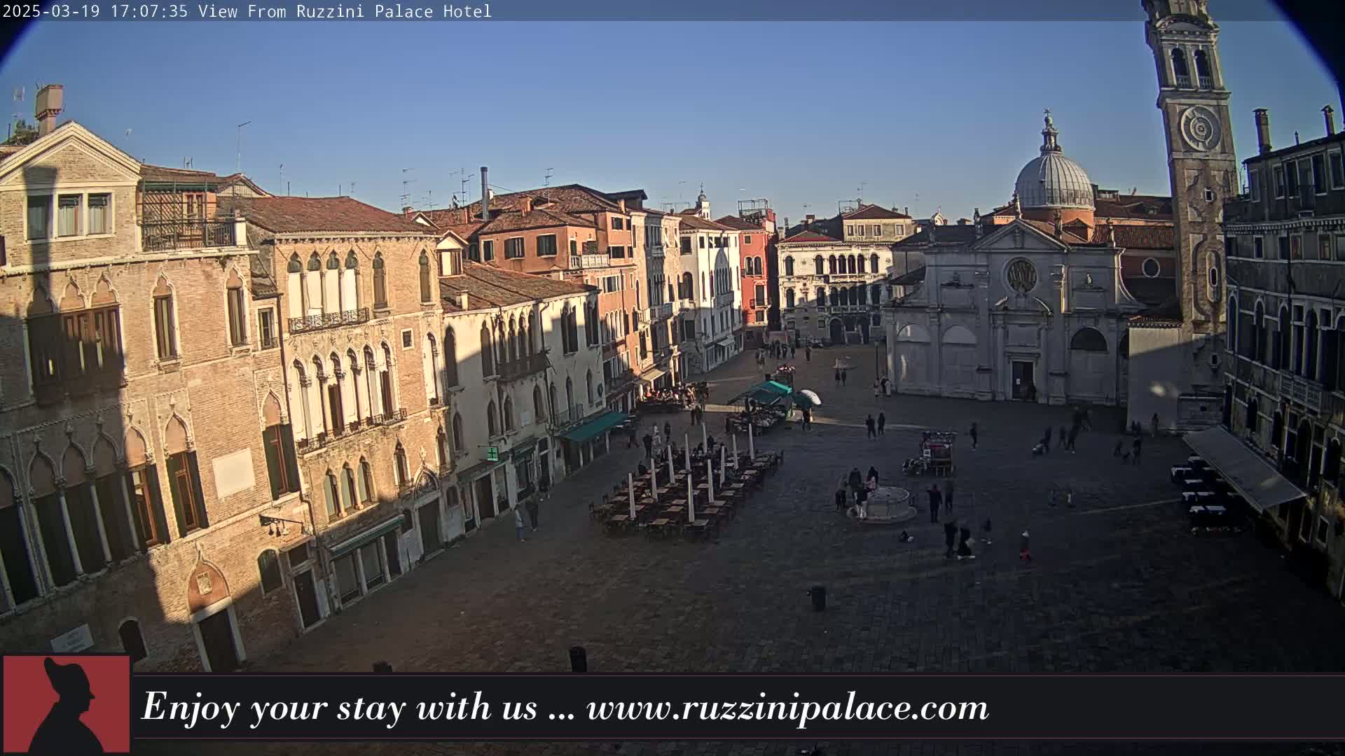 A sunny day reveals a large, mostly empty plaza in Venice, Italy, surrounded by historic buildings, with some people and outdoor seating visible.