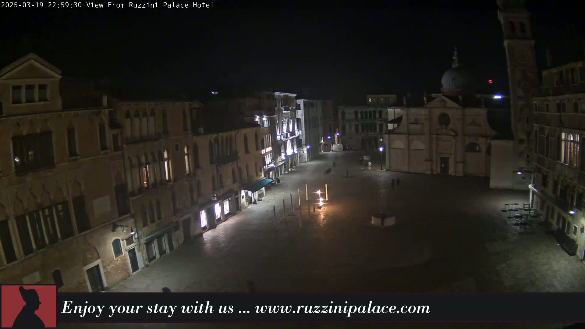 A nighttime view of a mostly empty plaza in Venice, Italy, surrounded by tall buildings and a church, under clear, dark skies.