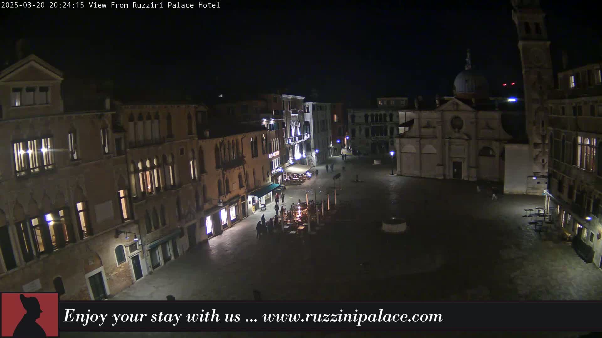 A nighttime, overhead view of a dimly lit plaza in Venice, surrounded by old buildings, with a few people visible and some outdoor seating.