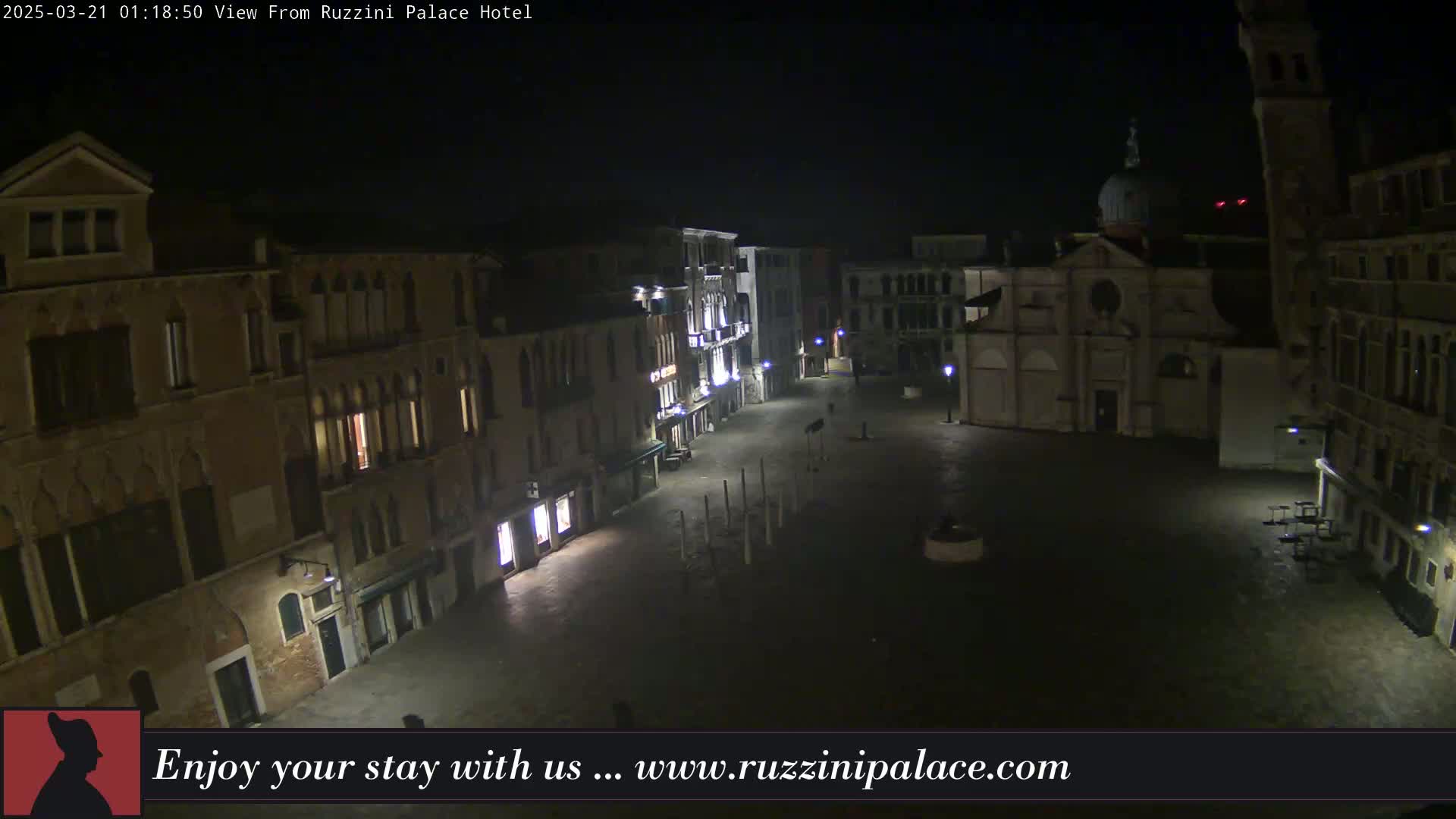 A nighttime view of a mostly empty square in Venice, Italy, surrounded by old buildings, with some outdoor seating visible.