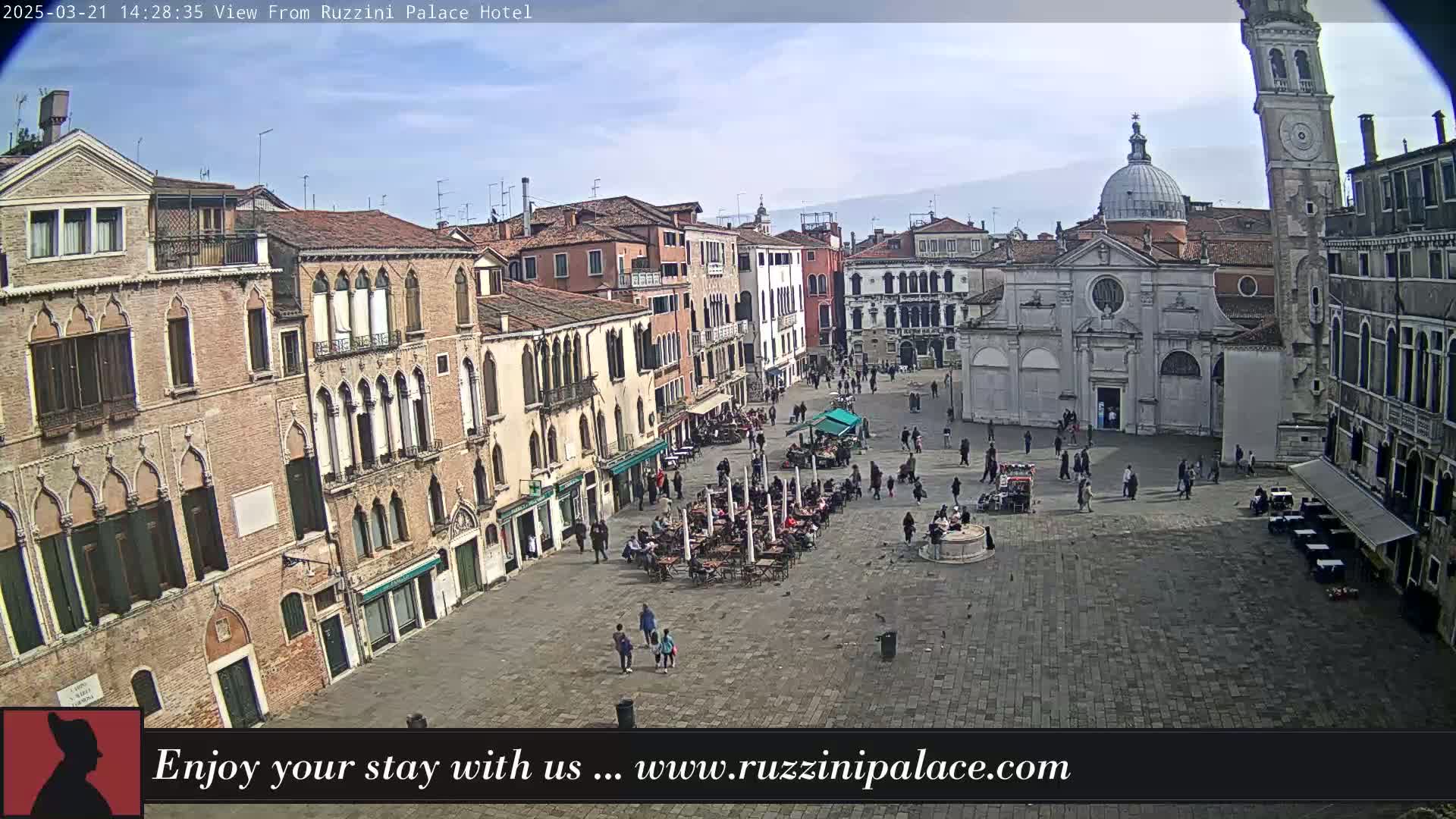 A mostly sunny day reveals a large, paved town square in Venice, Italy, surrounded by historic buildings, with people sitting at outdoor cafes and walking around.