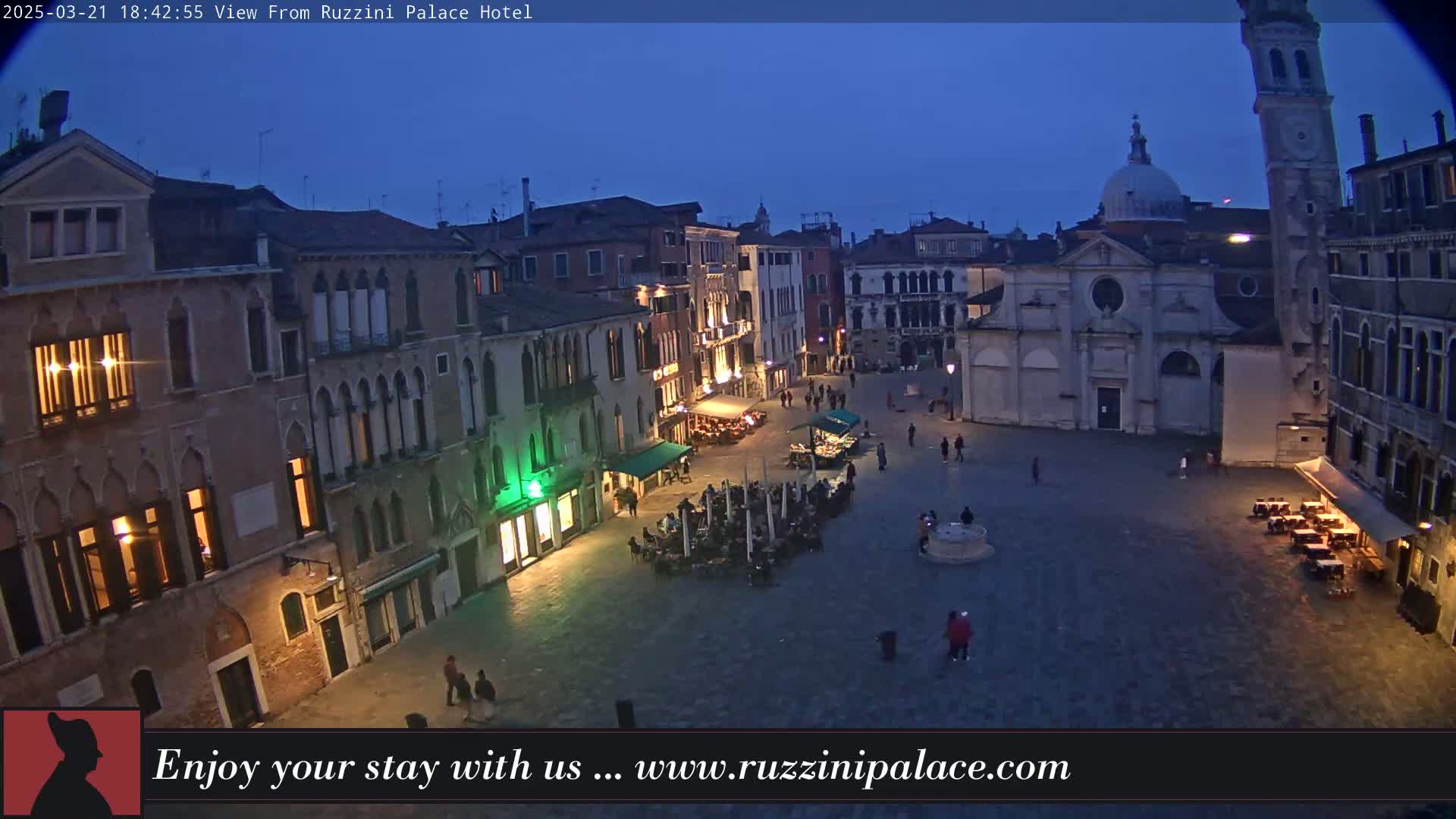 A twilight view of a large, mostly empty square in Venice, Italy, surrounded by old buildings, with a few people walking around and some outdoor seating at cafes.