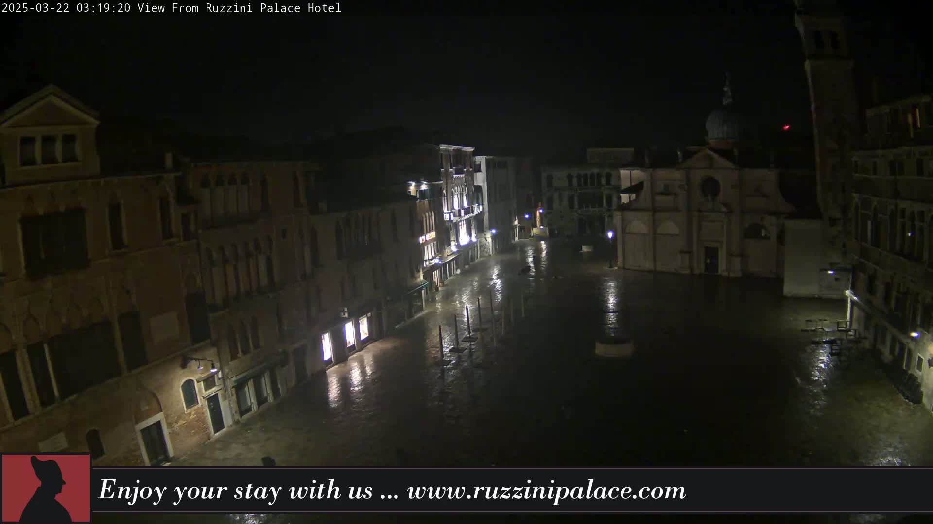 A nighttime view of a rain-soaked Venetian square, surrounded by old buildings, one of which is a church with a dome.