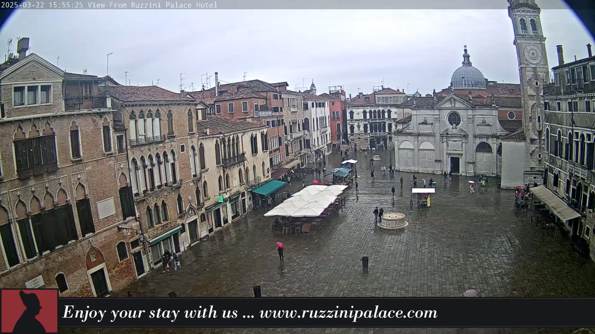 A rain-slicked Venetian piazza, surrounded by historic buildings, features a few people and outdoor seating under canopies.