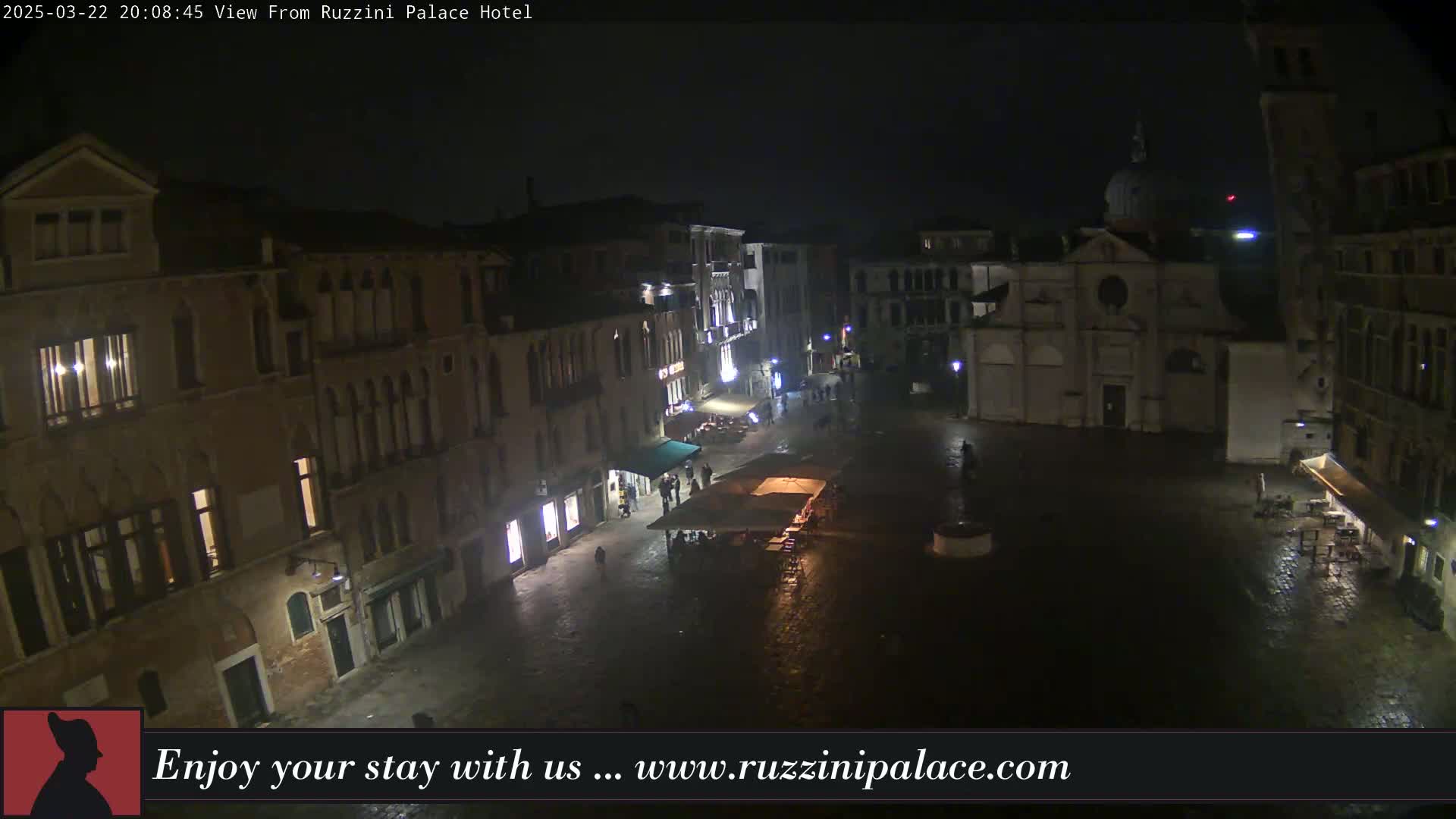 A nighttime view of a wet, dimly lit plaza in Venice, surrounded by buildings, with a few people and outdoor seating visible.