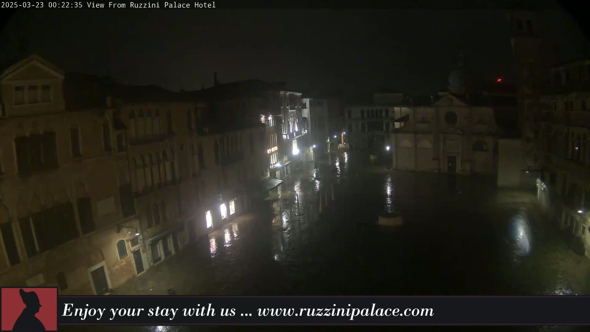 A nighttime, flooded Venetian square is seen from above, with buildings lining its sides and lit by streetlights.