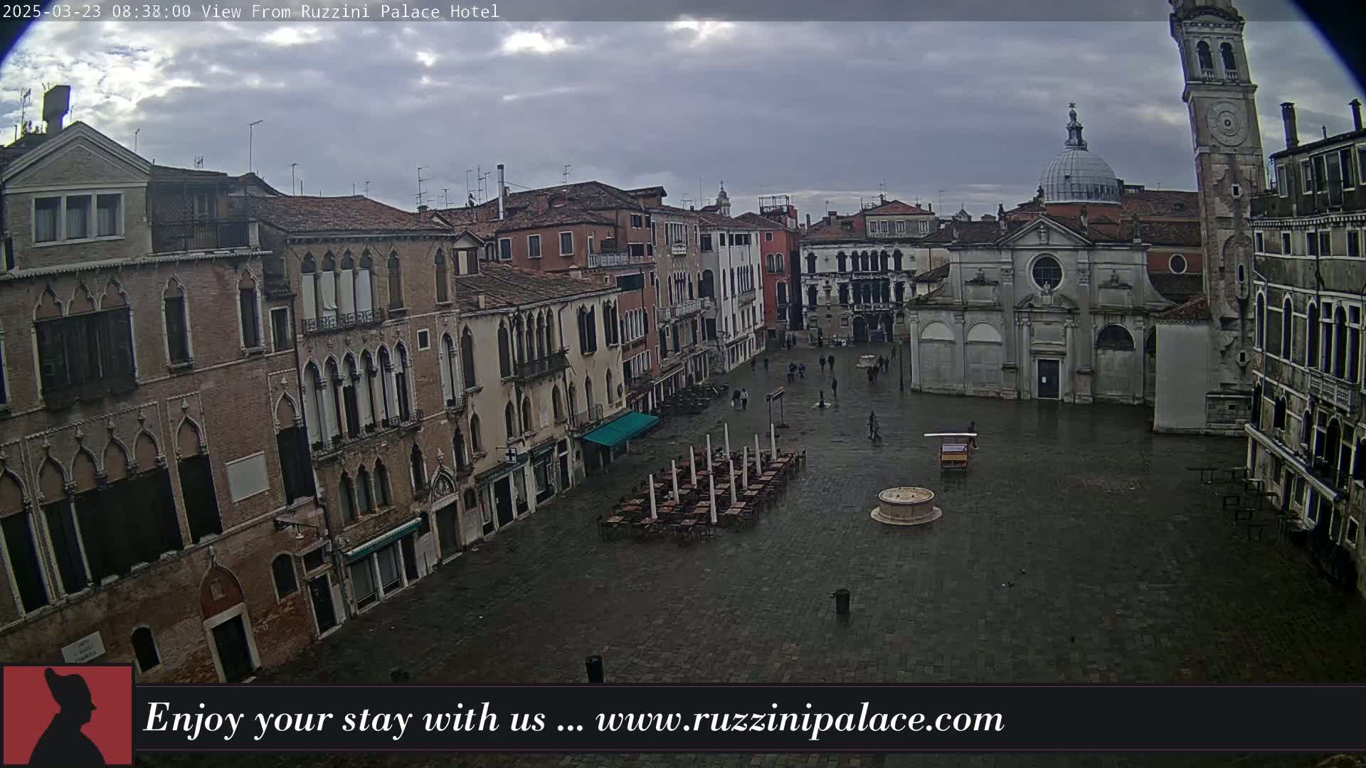 A wet, overcast square in Venice, Italy, is surrounded by historic buildings, with a few people and empty outdoor seating visible.