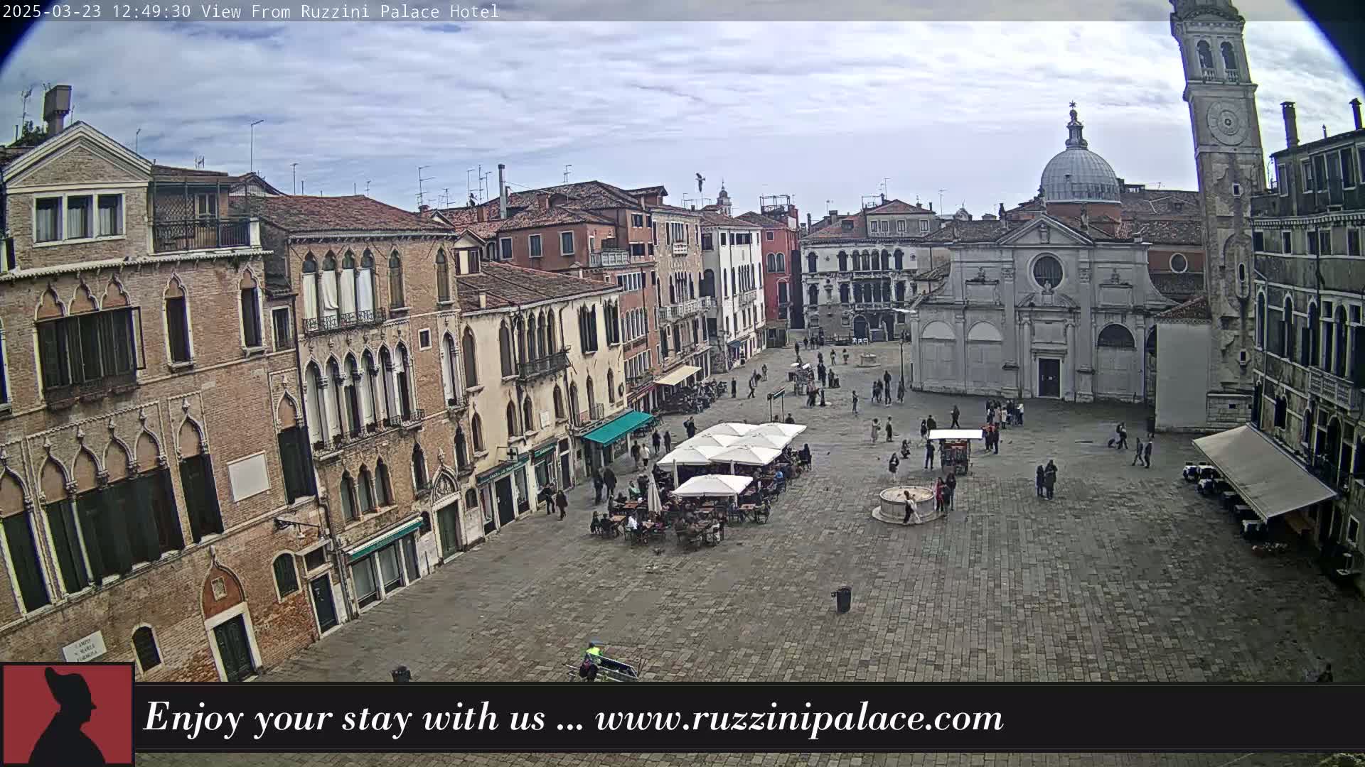 A large, stone plaza is surrounded by old, multi-story buildings under a mostly cloudy sky.
