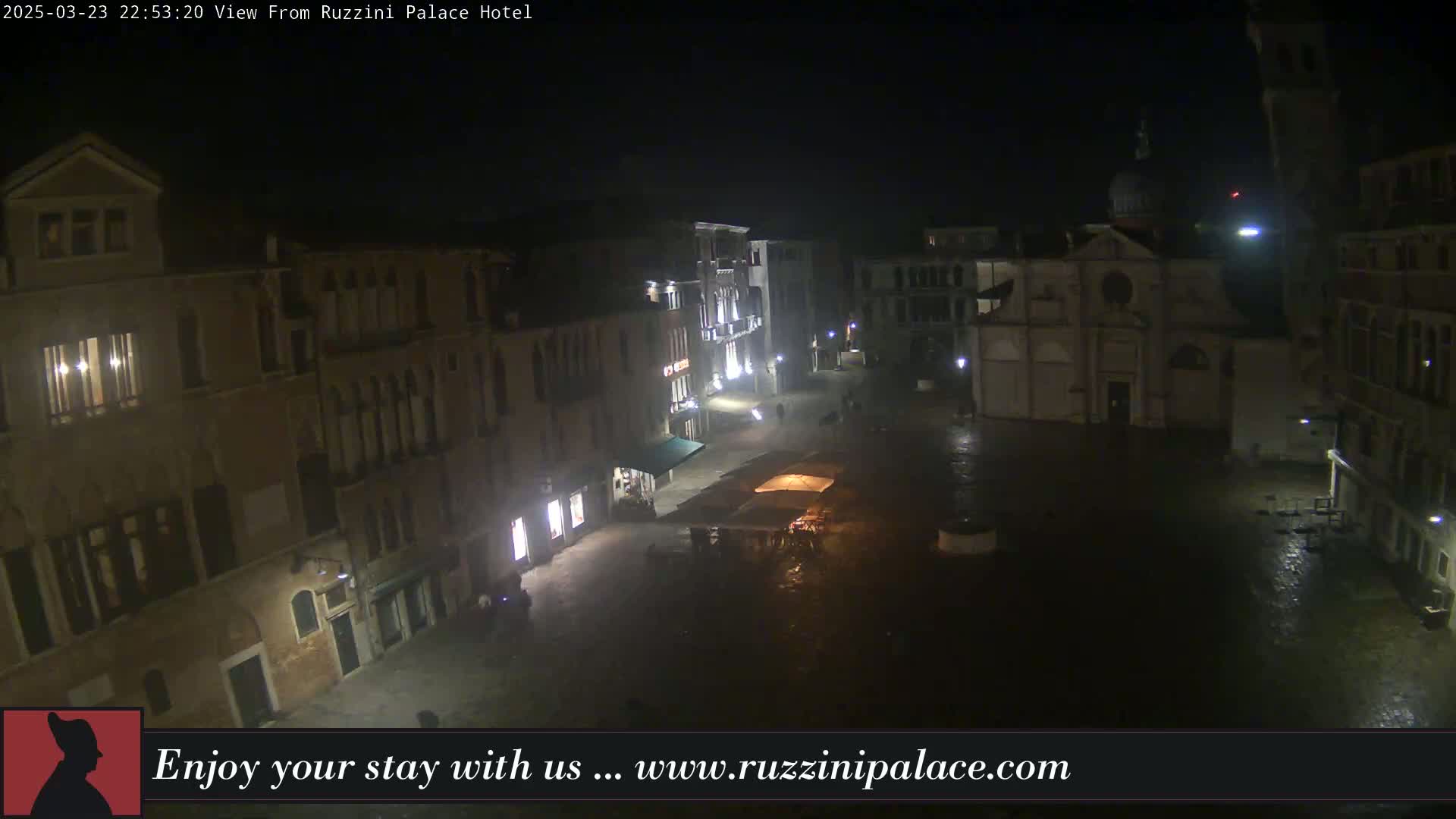 A nighttime, wet view of a dimly lit European town square, featuring several buildings, including a church.