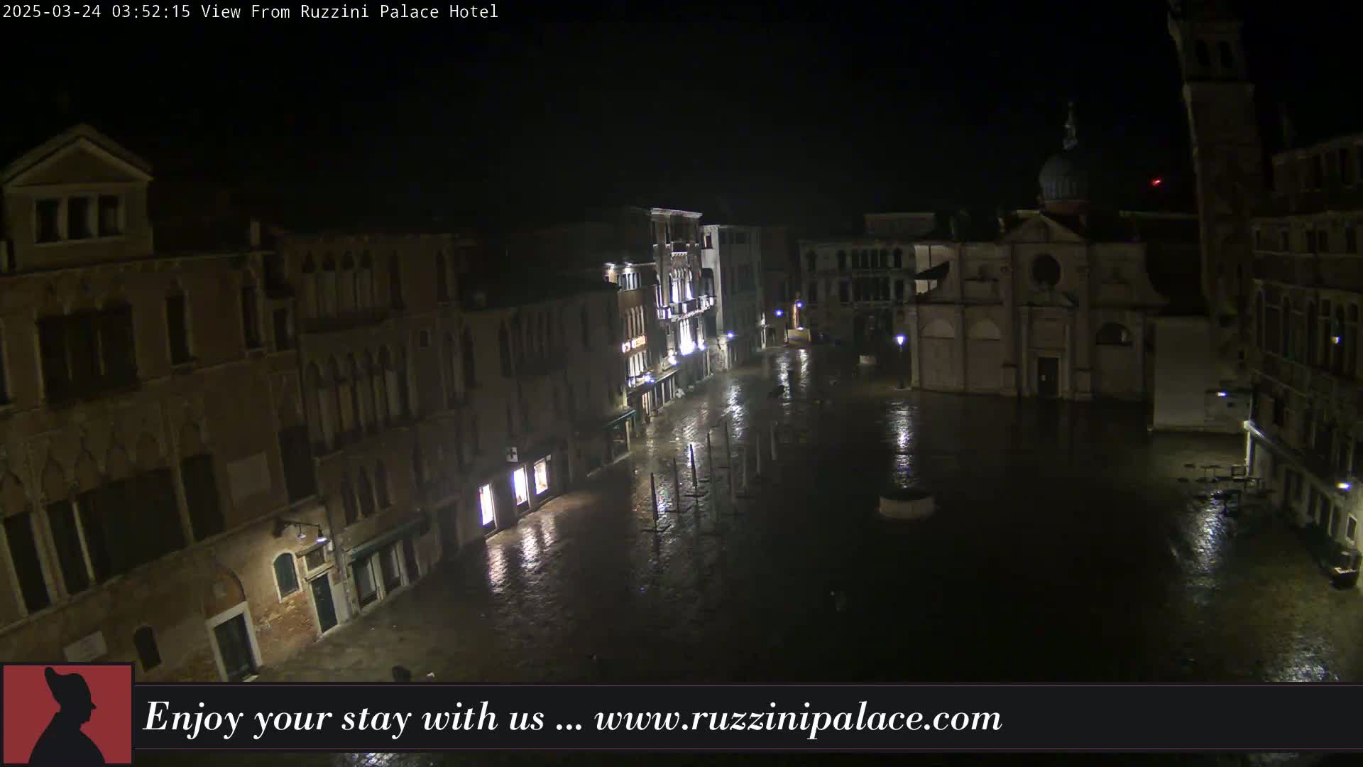 A nighttime, rain-soaked Venetian square is surrounded by illuminated buildings.