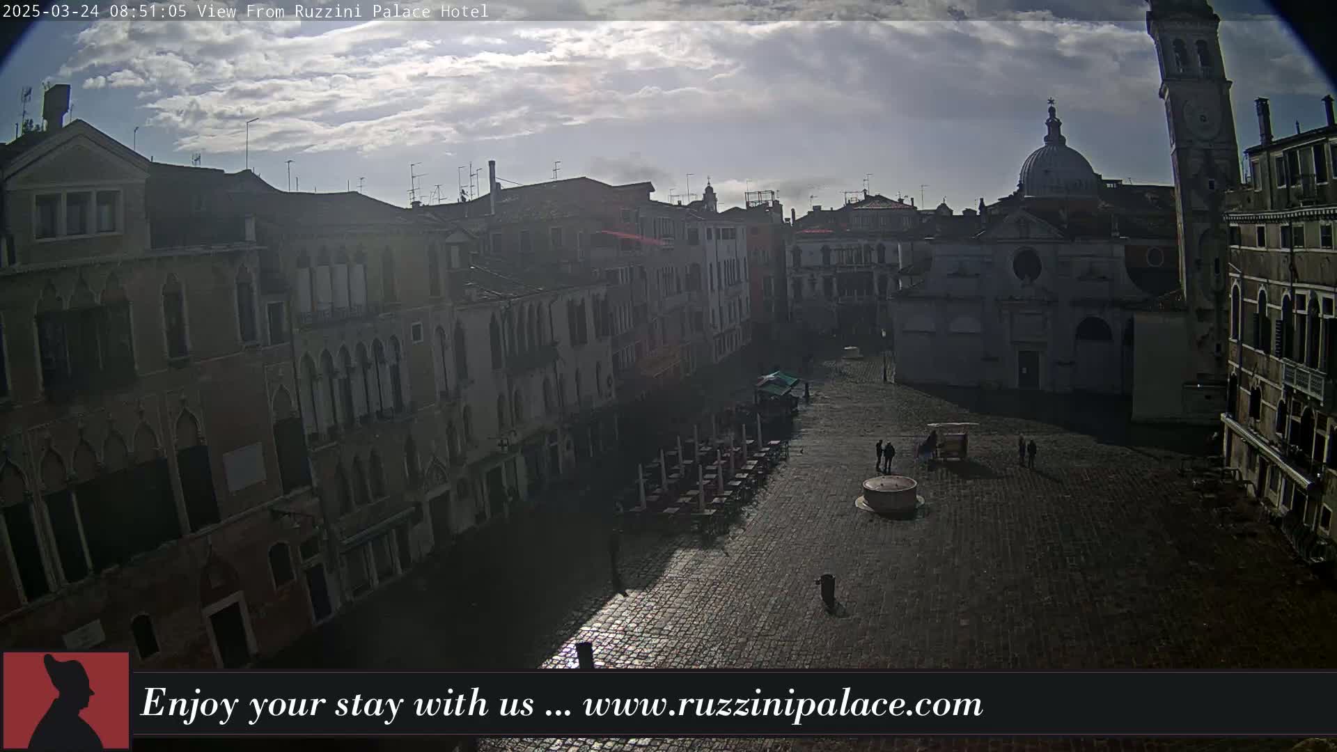 A mostly cloudy day reveals a cobblestone square in Venice, Italy, surrounded by historic buildings, with a few people and outdoor seating visible.