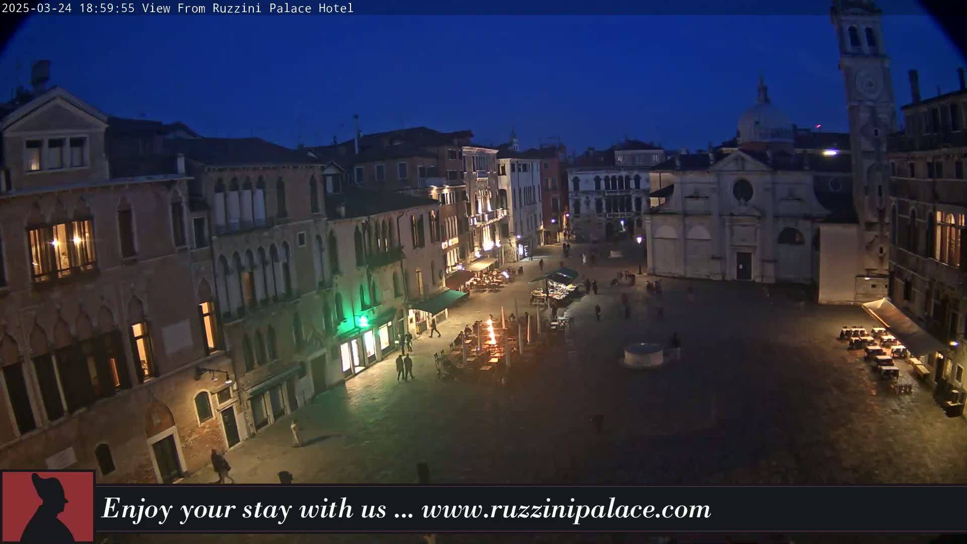 A nighttime view of a dimly lit European city square, surrounded by old buildings, with a few people and outdoor seating areas visible under a dark blue sky.
