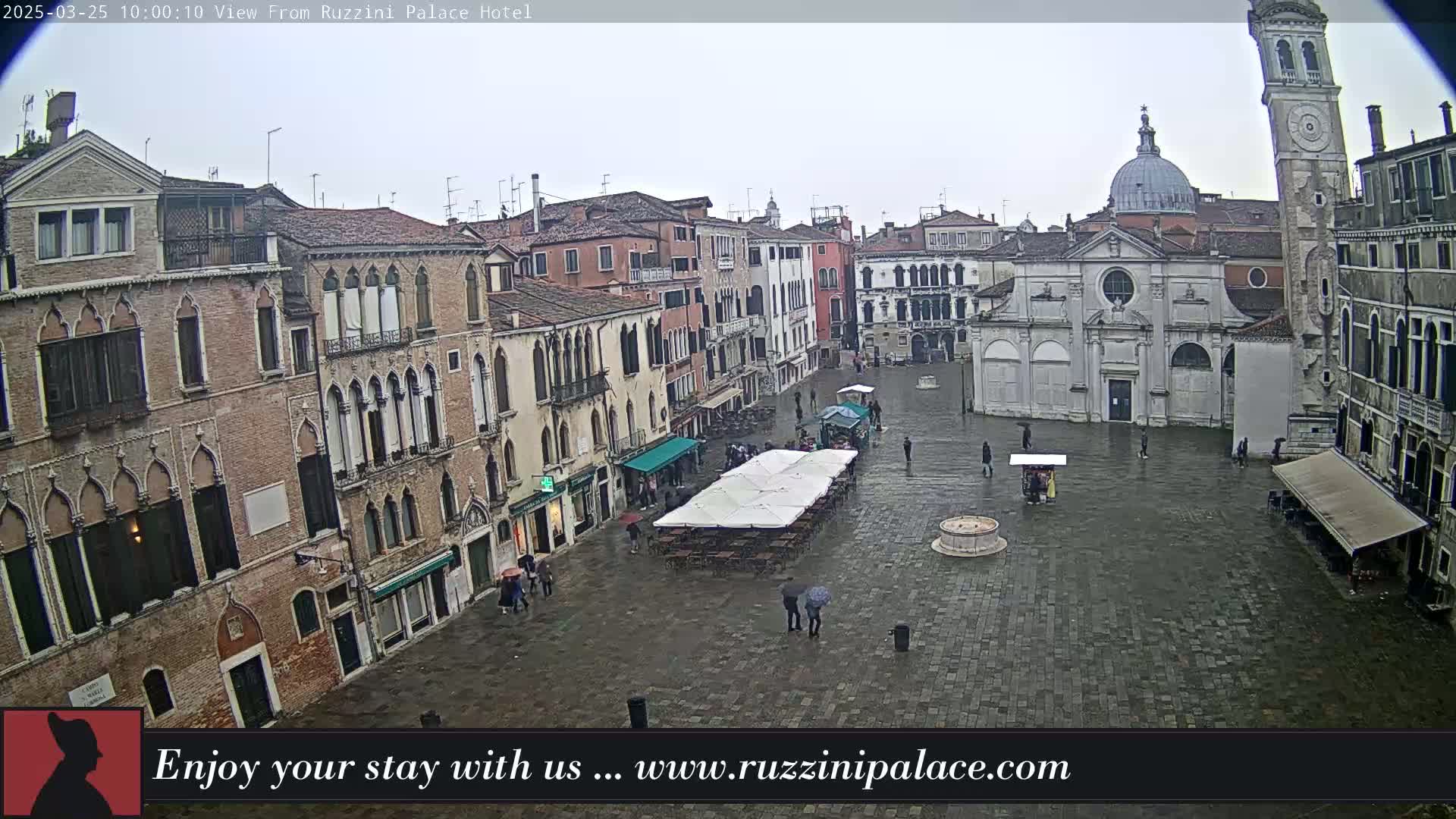 A rain-soaked Venetian square is viewed from above, showing several buildings, a church, and a few people walking amongst sparsely populated outdoor seating areas.