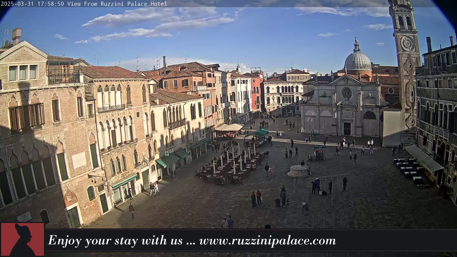 A sunny view of a large, paved square in Venice, Italy, surrounded by historic buildings, with several people and outdoor seating.