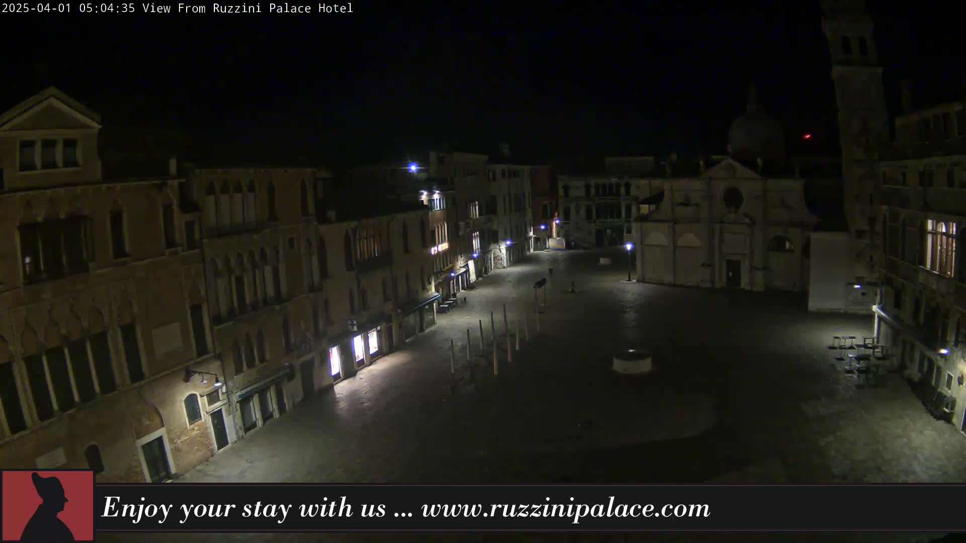 A nighttime, aerial view of a mostly empty, cobblestone plaza in a European city, surrounded by old buildings.