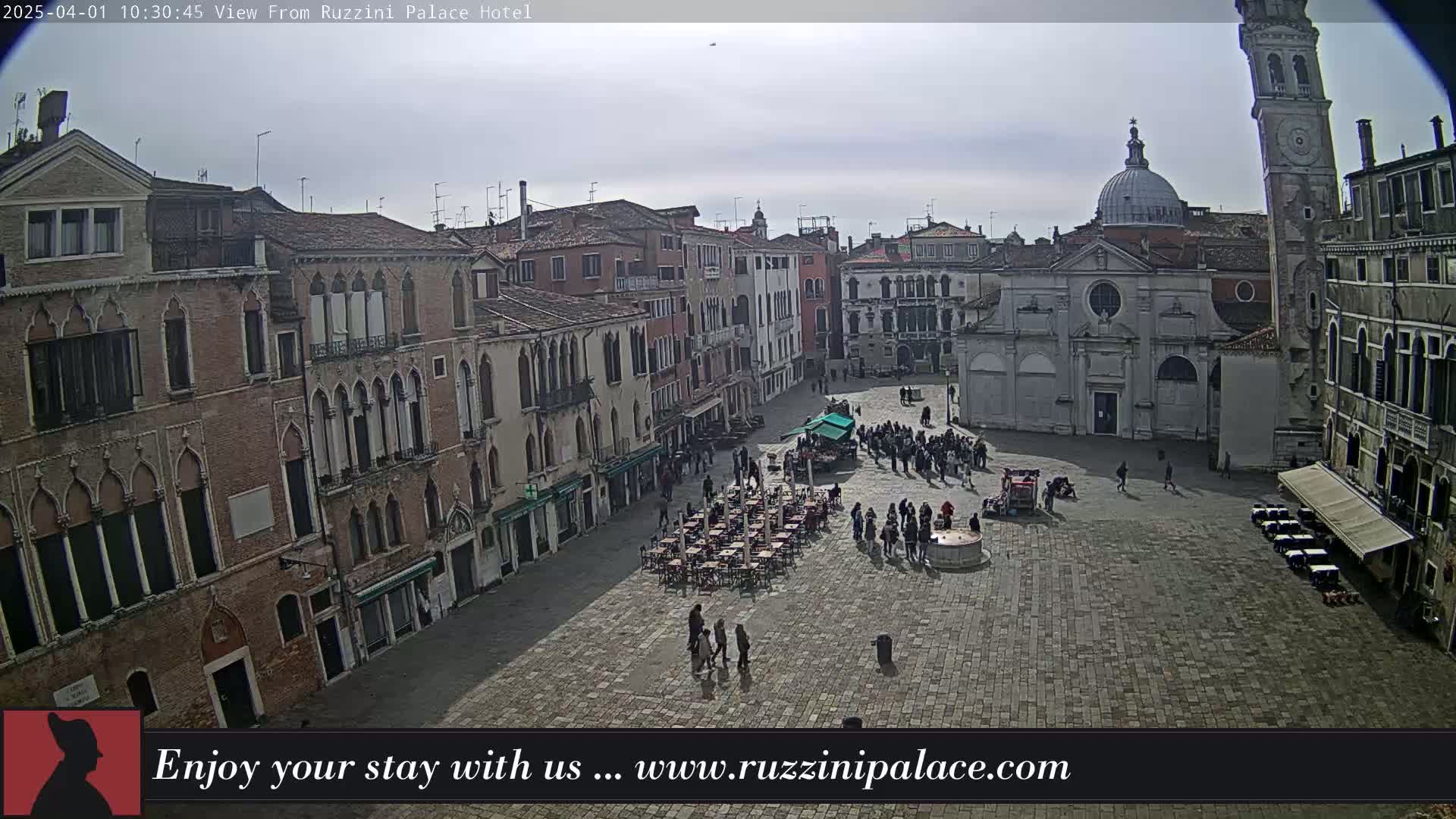 A mostly overcast day reveals a large, brick-paved square in Venice, surrounded by old buildings, with people walking and some seated at outdoor cafes.