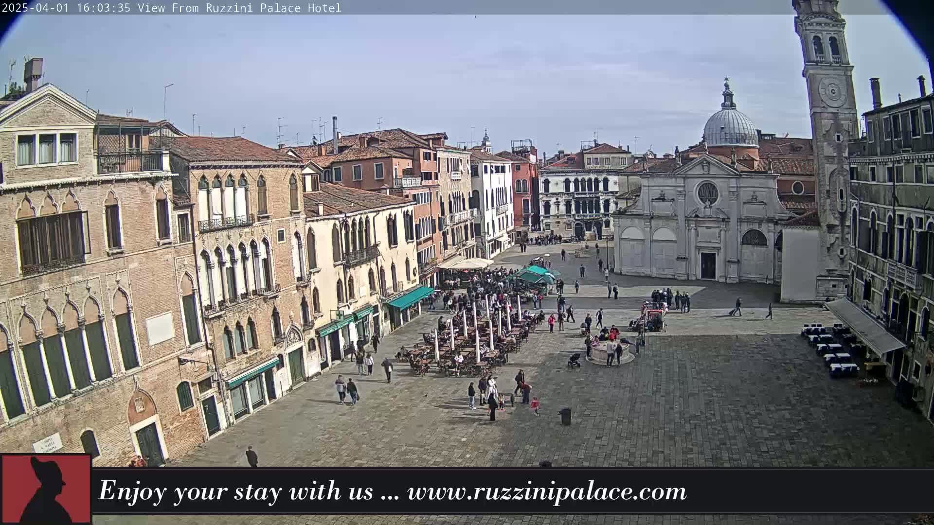 A sunny day in a large, paved European town square, filled with people and outdoor seating at cafes, surrounded by several-story buildings, one with a tall bell tower.