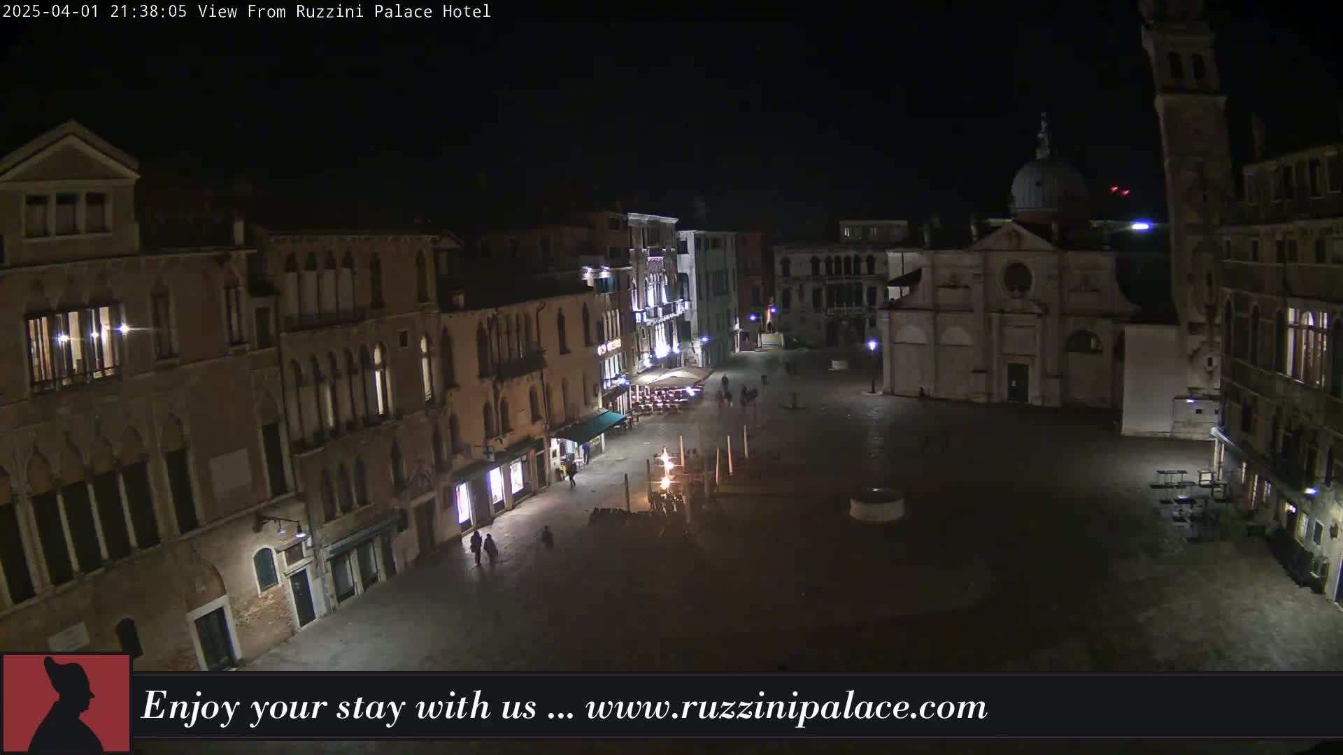A nighttime, high-angle view of a dimly lit, mostly empty European-style plaza surrounded by buildings.