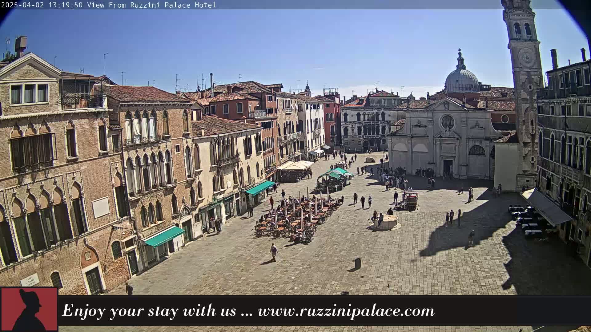 A sunny day reveals a large, cobblestone plaza in Venice, Italy, surrounded by historic buildings, with people sitting at outdoor cafes and strolling through the square.