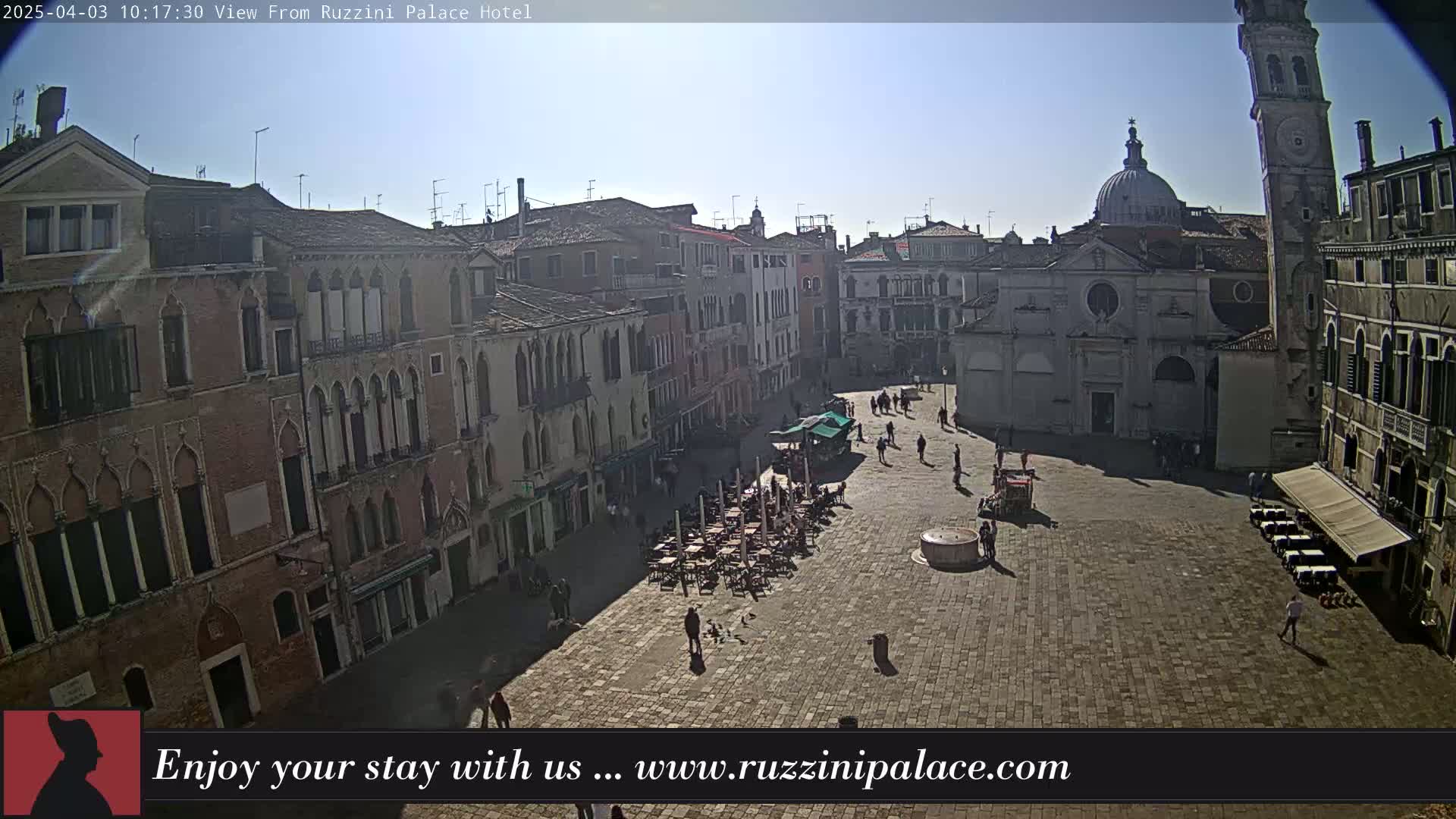 A sunny day reveals a large, paved square in Venice, Italy, surrounded by historic buildings, with a few people and outdoor seating visible.
