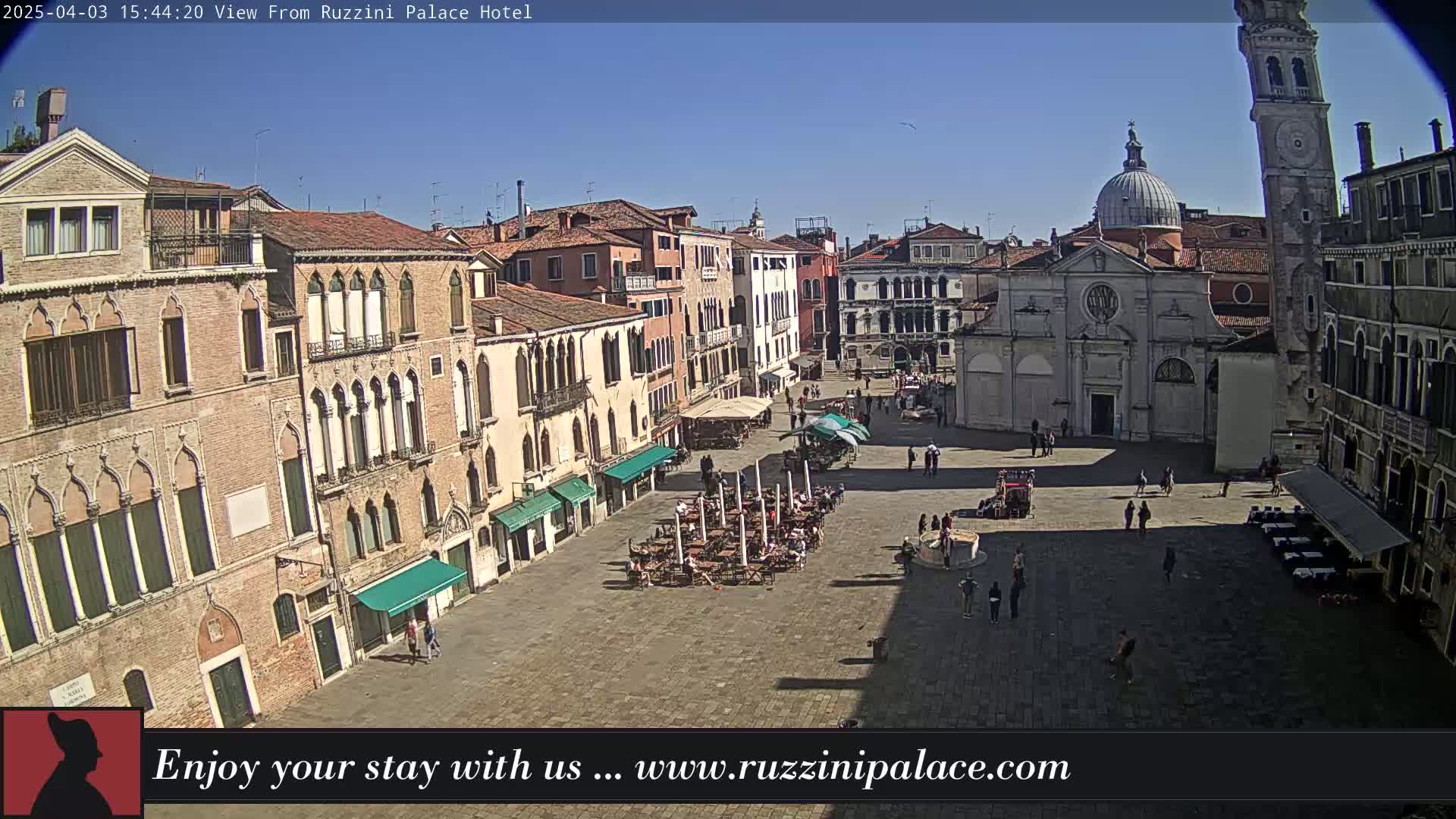 A sunny Venetian square is seen from above, showing several buildings, a church, a clock tower, and people scattered throughout the cobblestone plaza with outdoor seating at cafes.