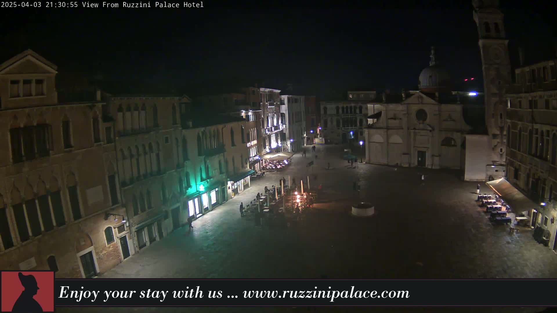 A nighttime, high-angle view of a dimly lit European plaza, surrounded by old buildings, with a few people and outdoor seating areas visible.