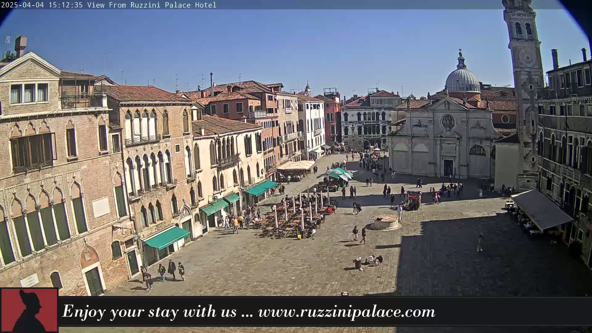 A sunny, outdoor view of a large plaza in Venice, Italy, surrounded by historic buildings, with people walking and sitting at outdoor cafes.