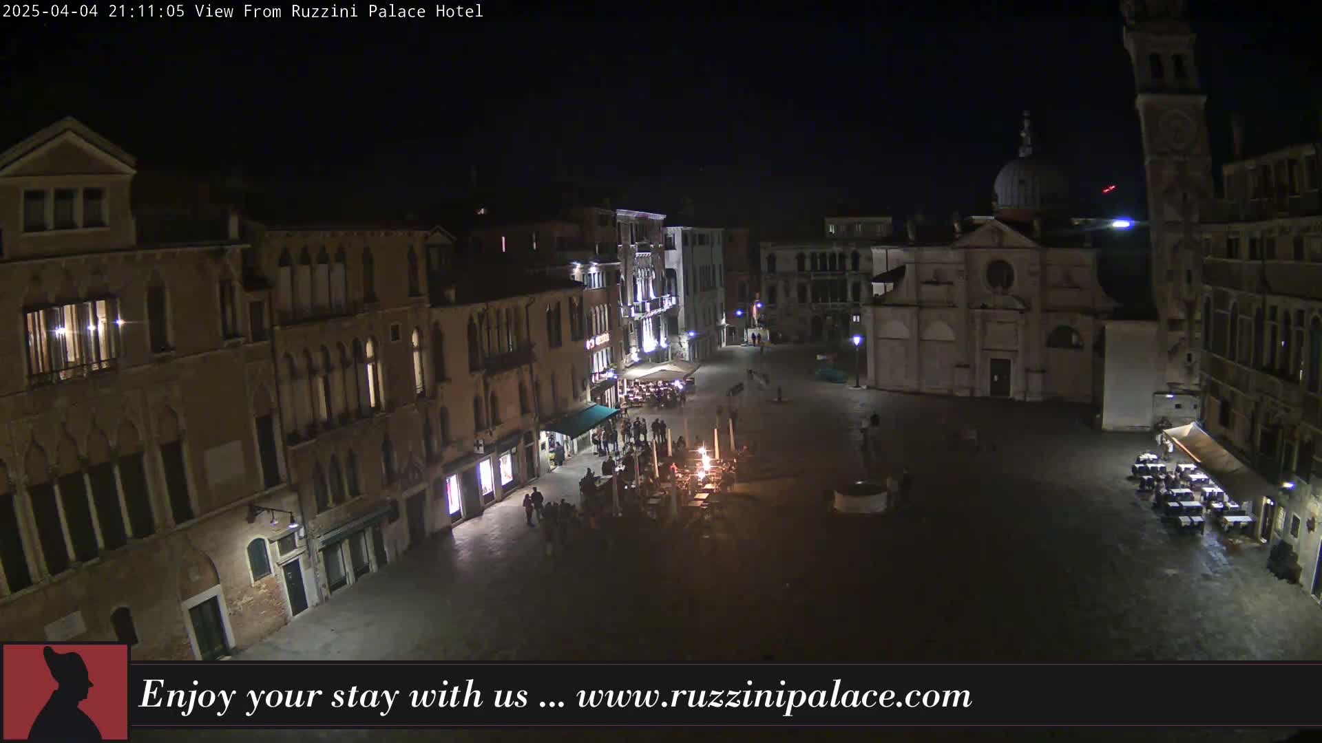 A nighttime, high-angle view shows a dimly lit European square with several buildings, some outdoor seating, and a small gathering of people.