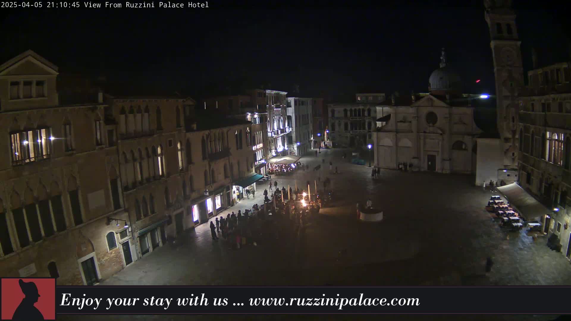 A nighttime, aerial view of a dimly lit European-style plaza with several buildings surrounding it, some with lights on in their windows, and a few people gathered in the square.