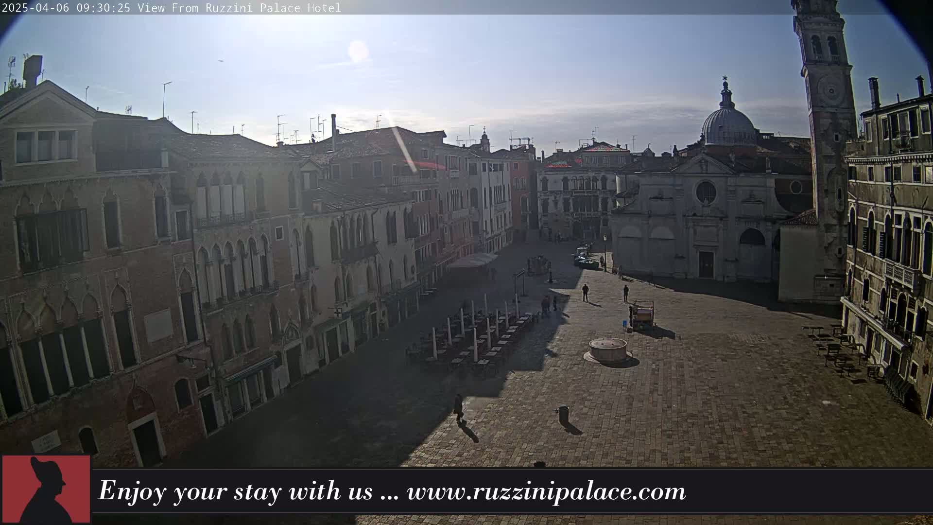 A sunny day reveals a large, empty plaza in Venice, surrounded by historic buildings, with a few people walking and some outdoor furniture set up.