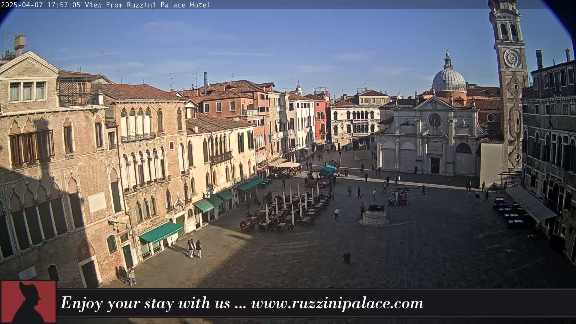 A sunny view of a large, paved square in Venice, Italy, surrounded by historic buildings, with outdoor seating and a few people scattered throughout.