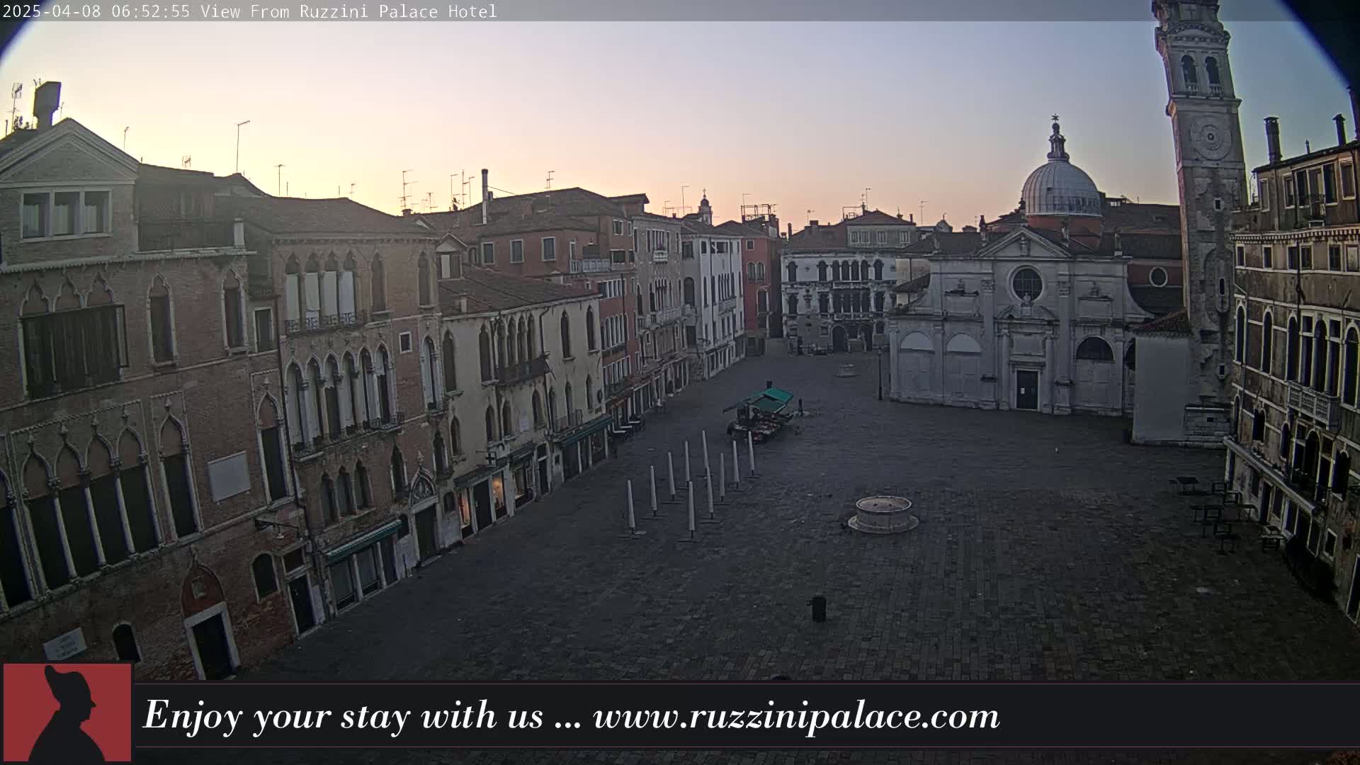 A wide, empty square in a city with several brick buildings, a church, and a tall tower, at sunrise.