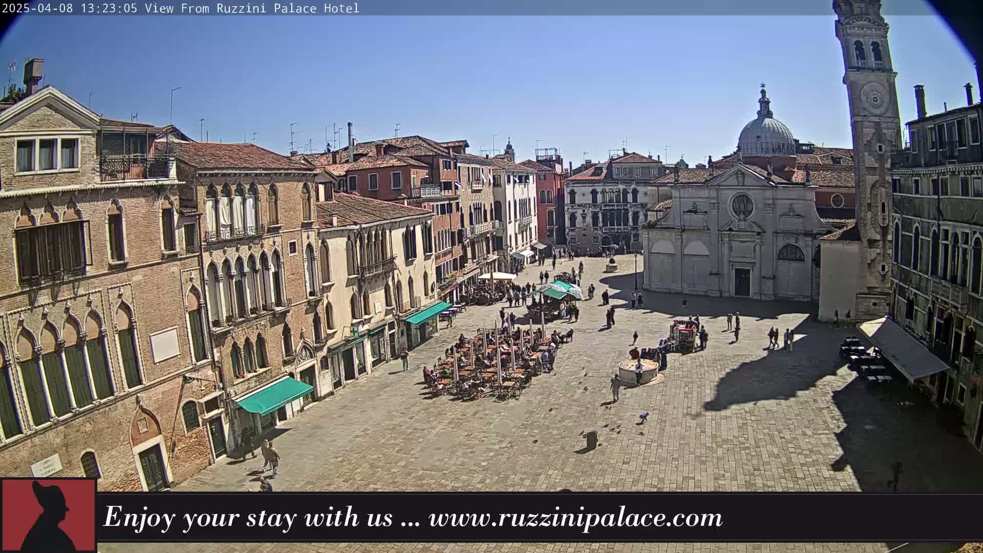 A sunny Venetian square is filled with people sitting at outdoor cafes, with several buildings surrounding it, including a church and a tall clock tower.