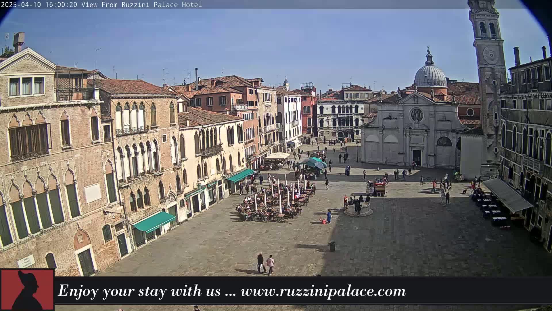 A sunny, outdoor view of a large plaza in Venice, Italy, surrounded by historic buildings, with people walking and sitting at outdoor cafes.