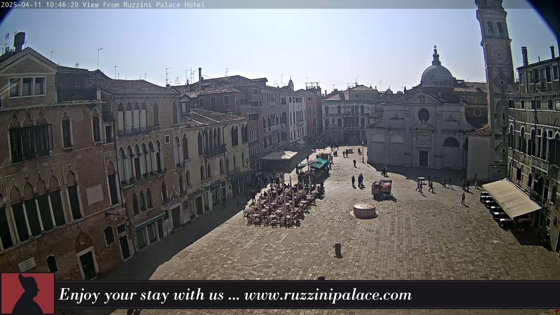 A sunny day reveals a large, paved square in Venice, surrounded by historic buildings, with a few people walking and some outdoor seating at cafes.